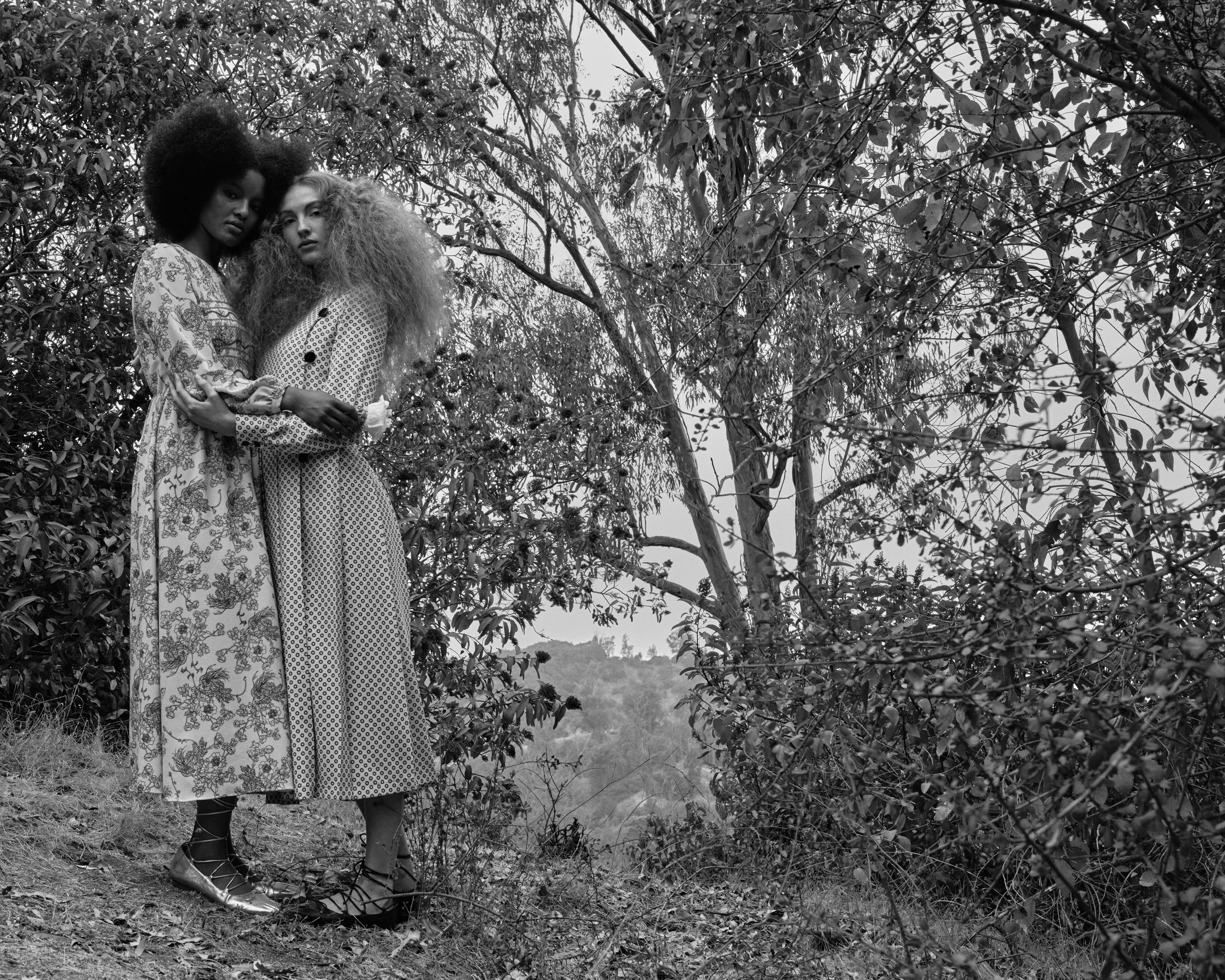 Two women with voluminous curly hair standing close together outdoors amidst trees and foliage, wearing patterned dresses, one in floral and the other in geometric print, holding each other's arms, in a black and white photograph.