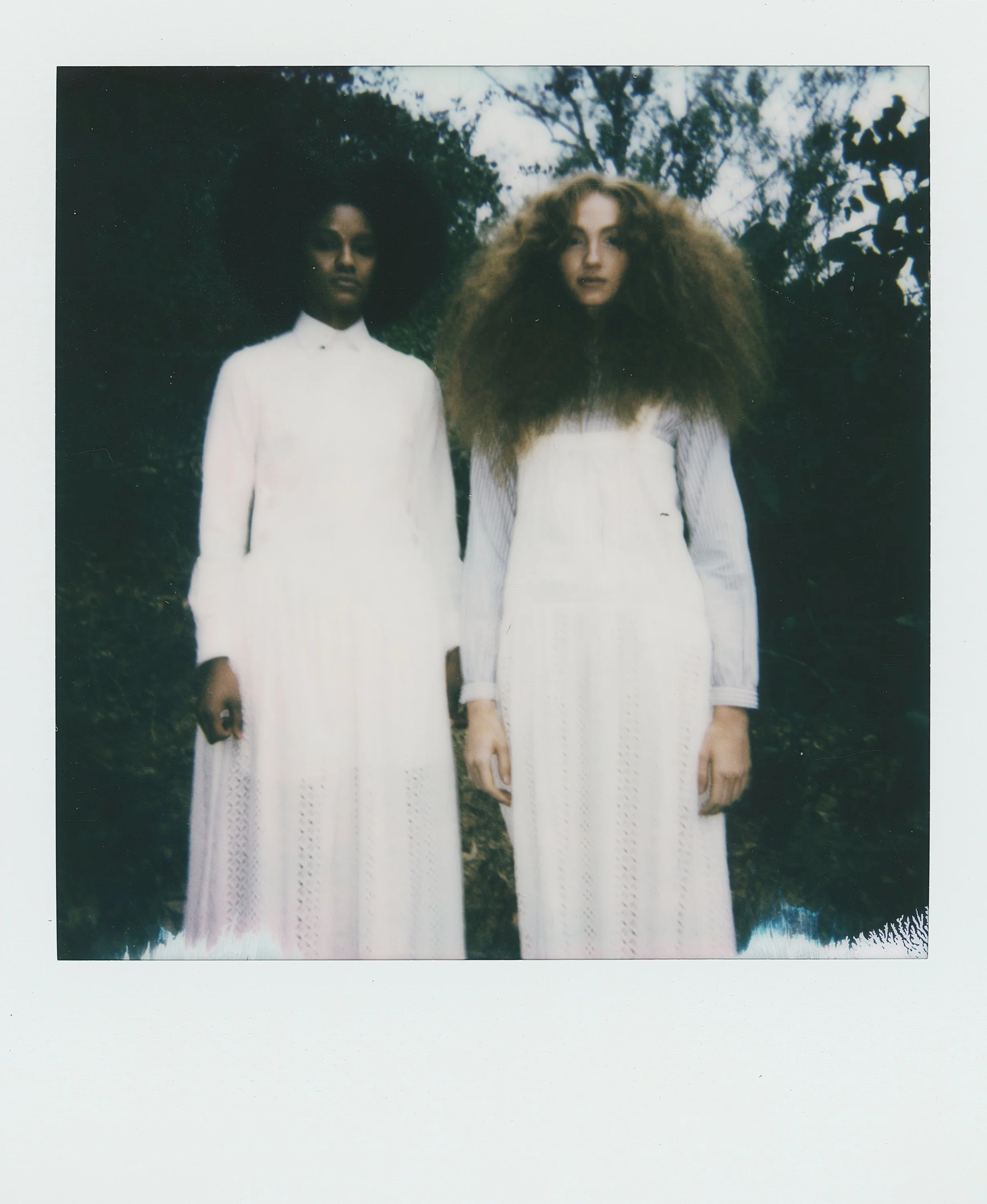 Two women standing outdoors in front of trees, wearing long white dresses with intricate lace details, with voluminous curly hair.