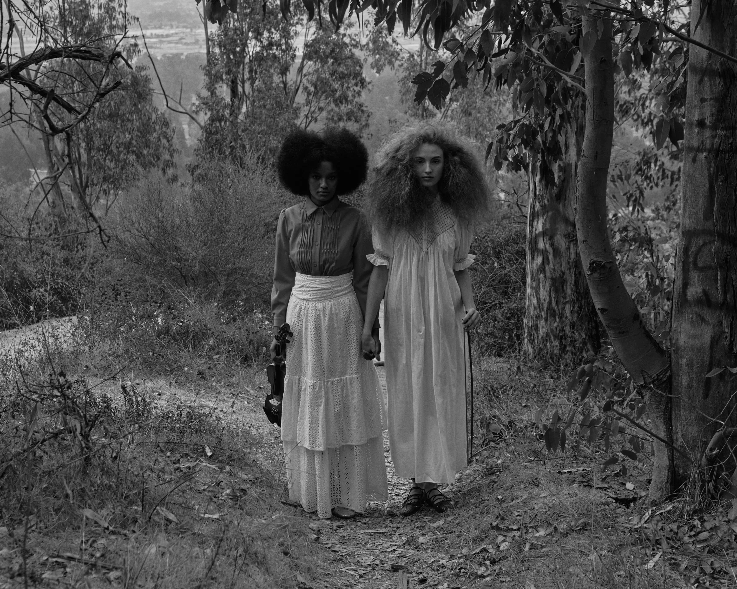 Two women with large curly hair walking through a wooded area, holding hands, in black and white.