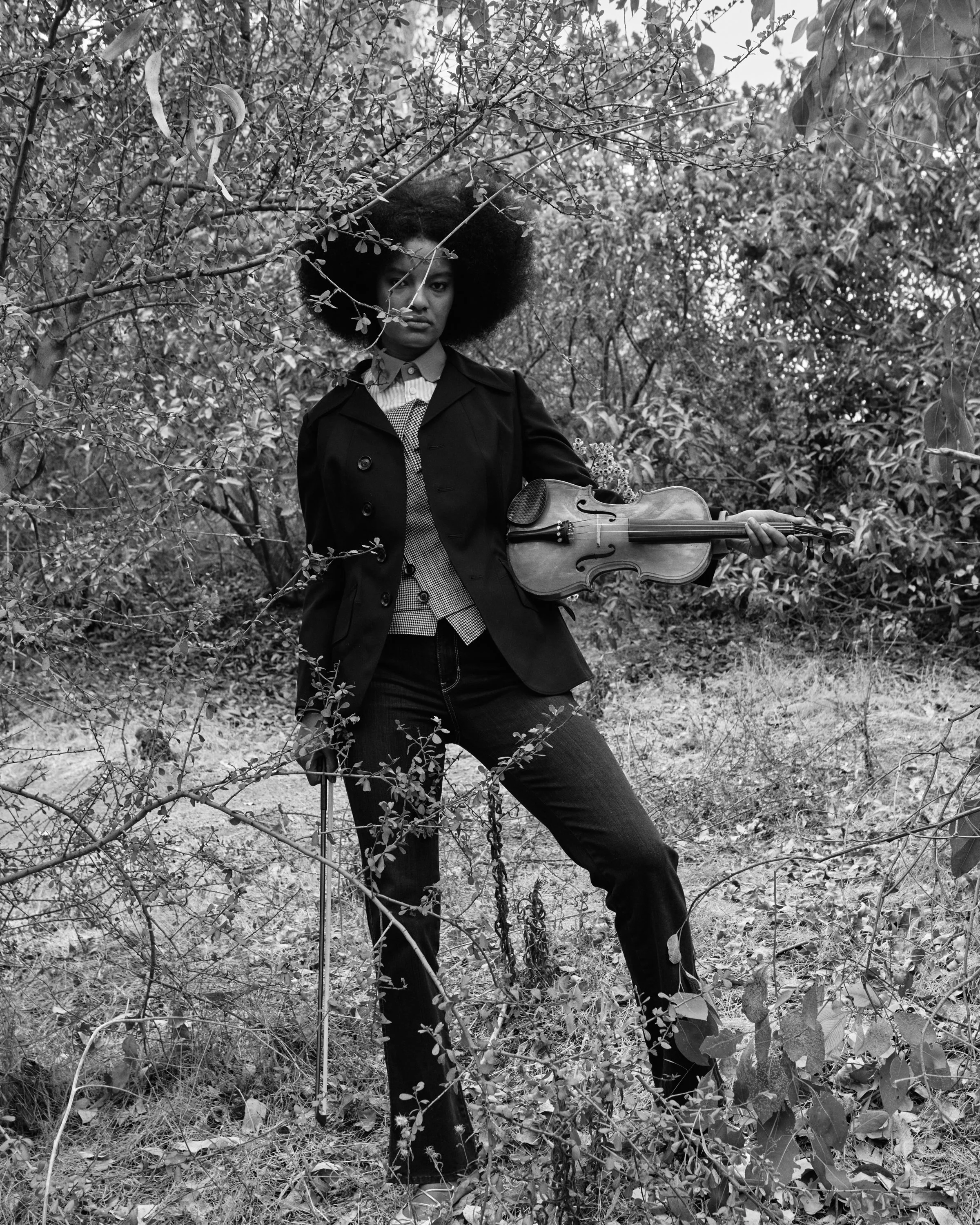 A woman standing outdoors among bushes, holding a violin and a bow, with an afro hairstyle, dressed in dark pants and a jacket.