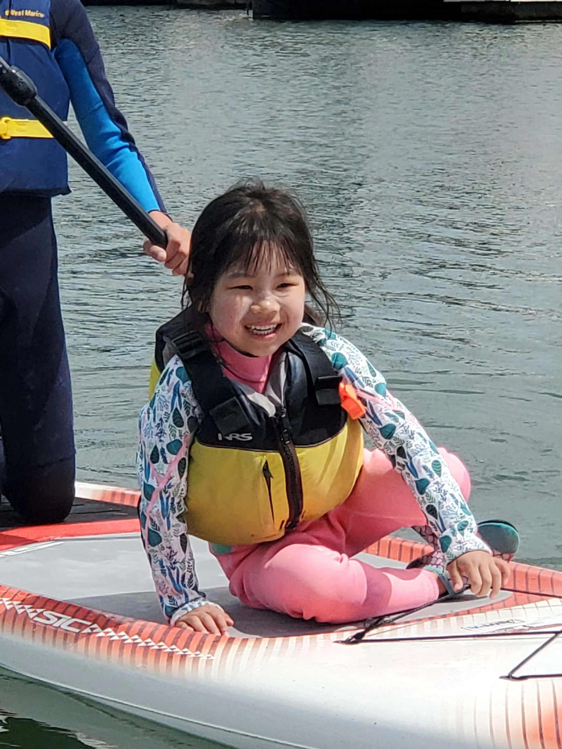 A young girl in a pink outfit and floral jacket sits on a paddleboard, smiling cheerfully, enjoying the best Bay Area summer camp.