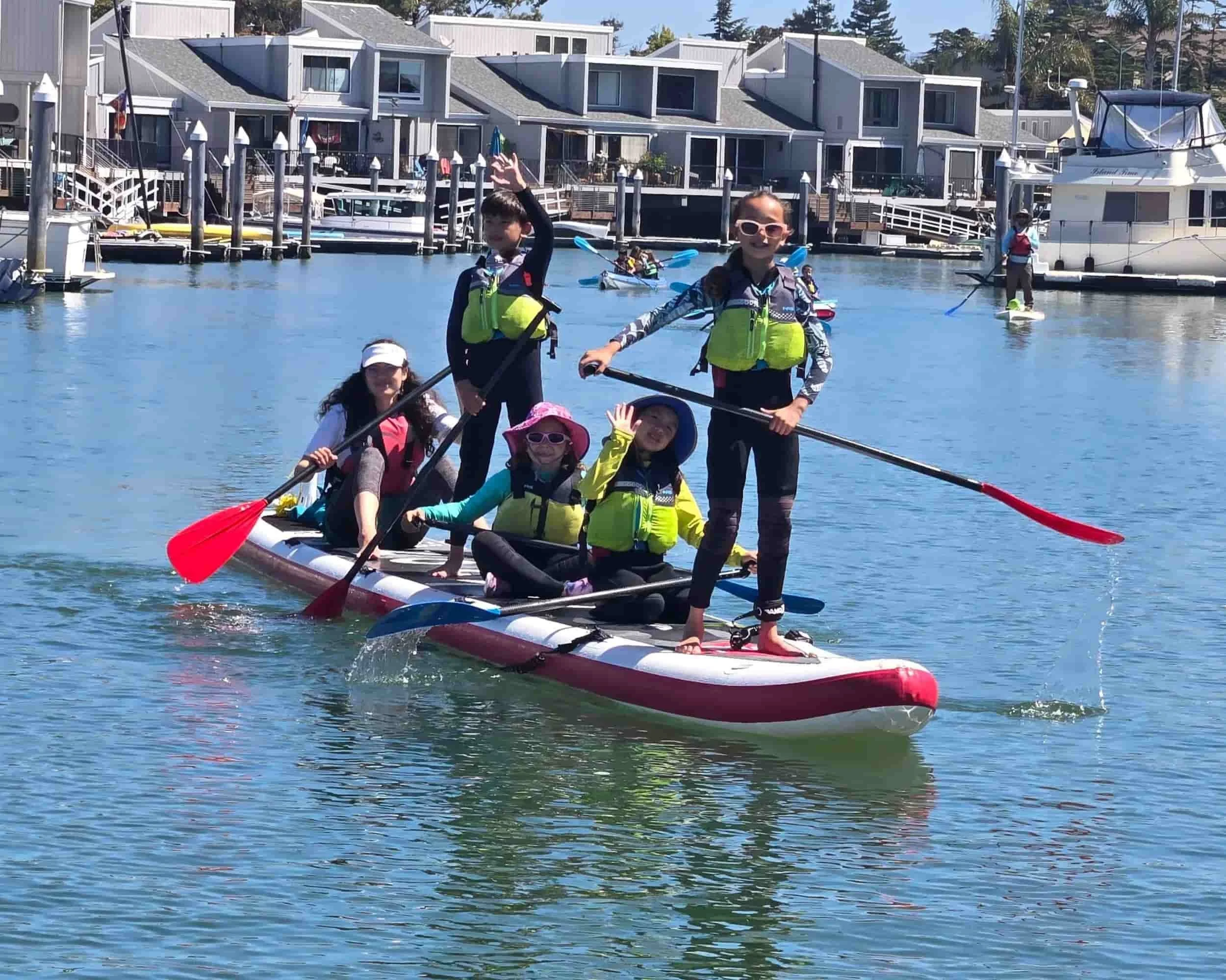 Group of five children on a stand-up paddleboard, wearing life jackets, for summer adventure camp in Alameda, CA