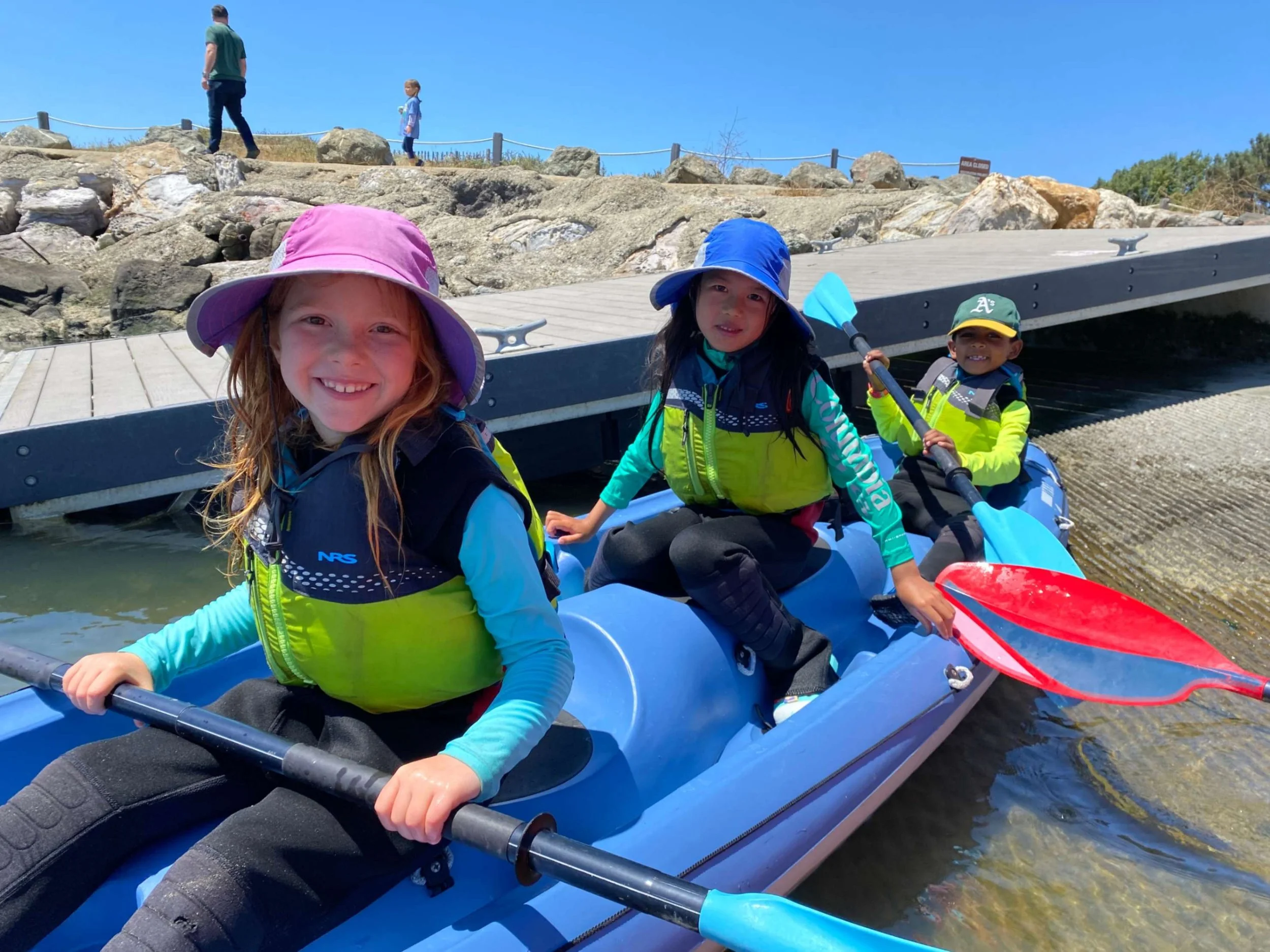 Three children in life jackets and sun hats paddling in a blue kayak on a sunny day, for the best East Bay summer camp.