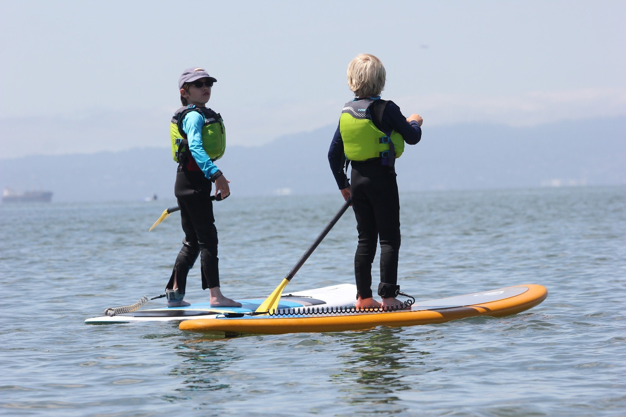 Two children stand on paddleboards on a calm body of water, on the San Francisco Bay for summer camp.