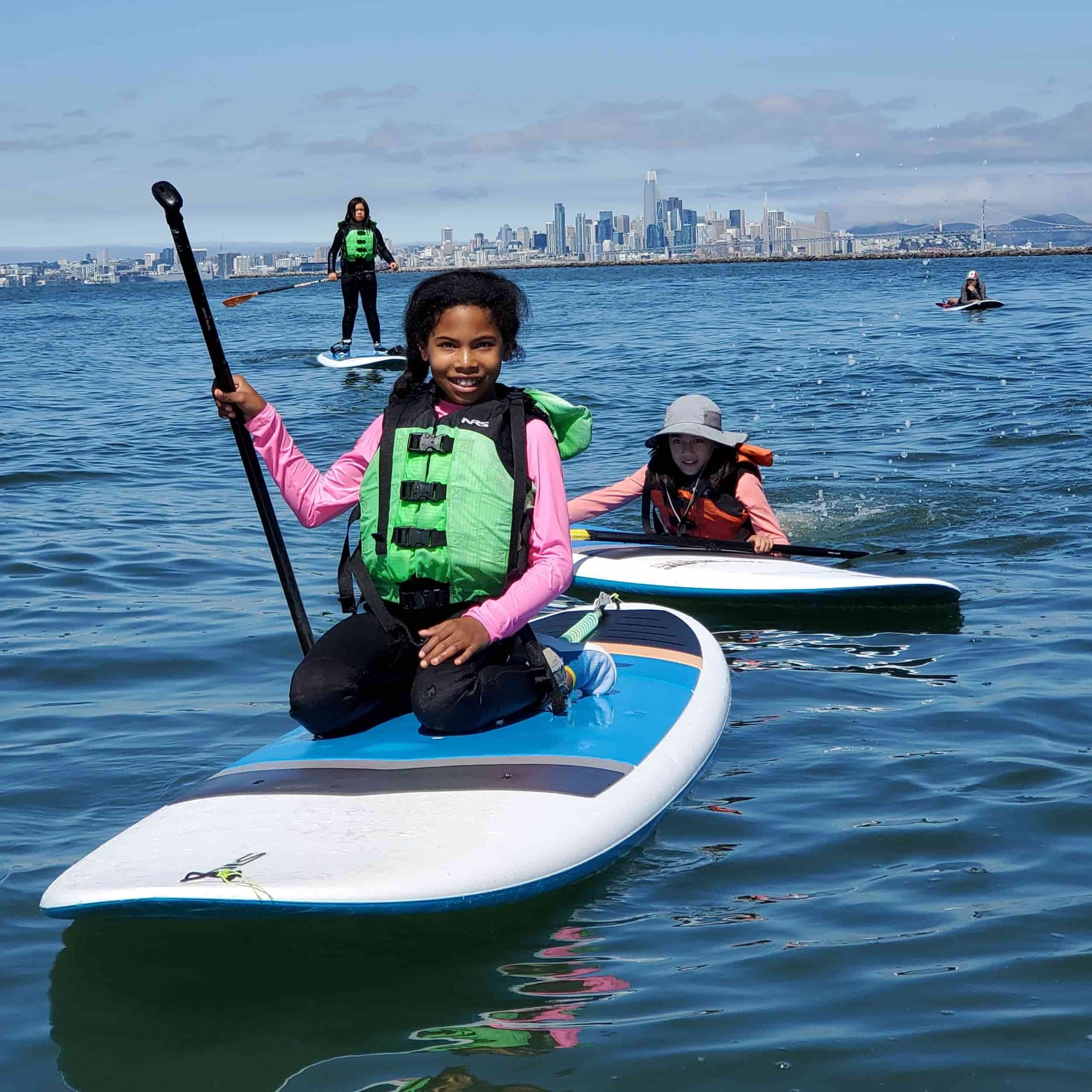 Children paddleboarding on the water with the San Francisco skyline in the background.