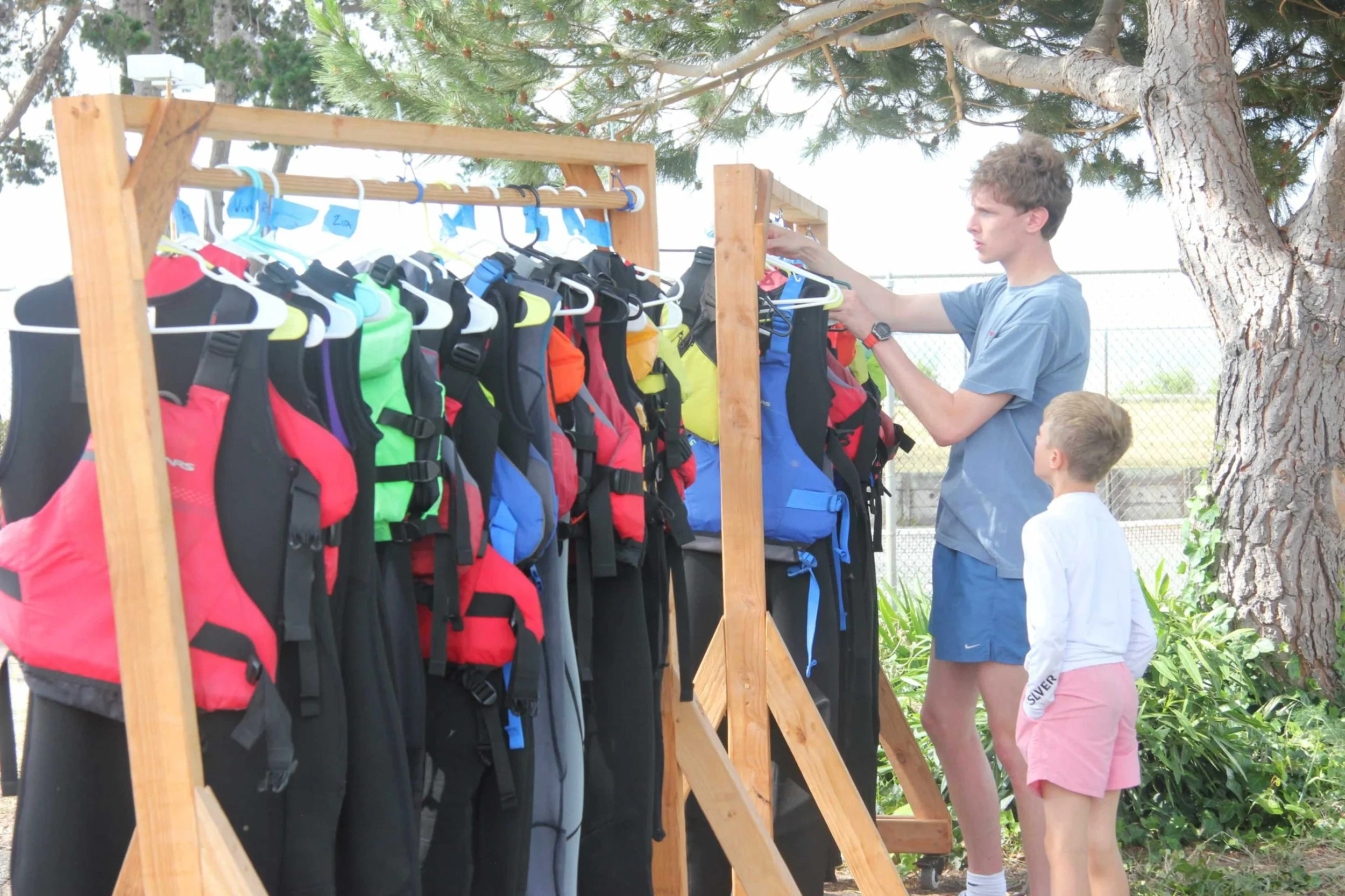 A young man and boy are looking at life jackets and wetsuits hanging on a wooden rack for the best bay areas summer camp.