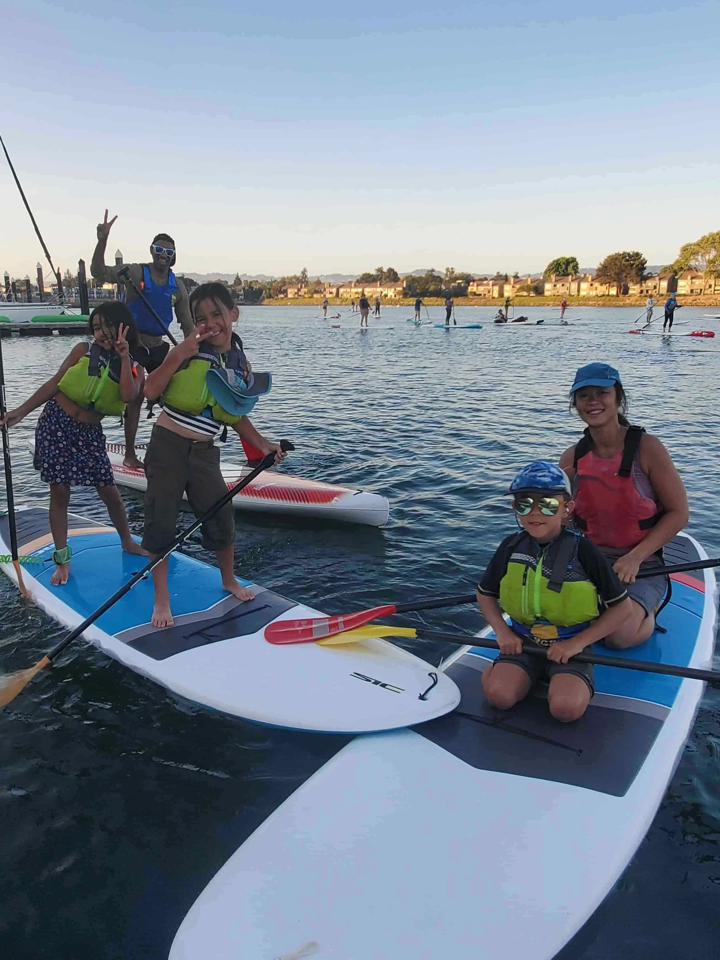A Bay Area family on paddleboarding on a calm body of water during sunset, for Family Paddle Nights.