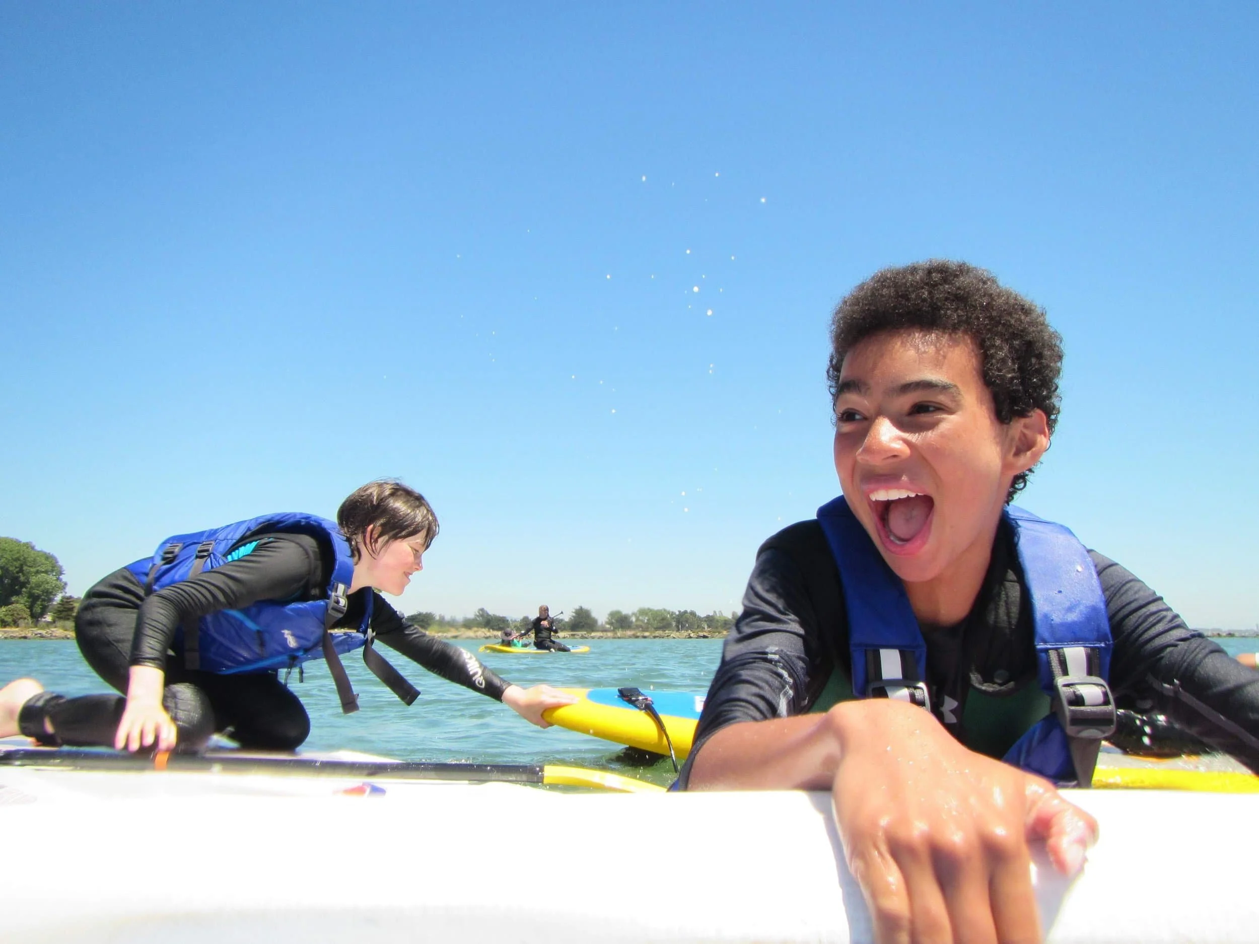 Two children on paddleboards, one smiling and grabbing the edge, the other kneeling and reaching for an item on the board, at the best summer camp in the East Bay.