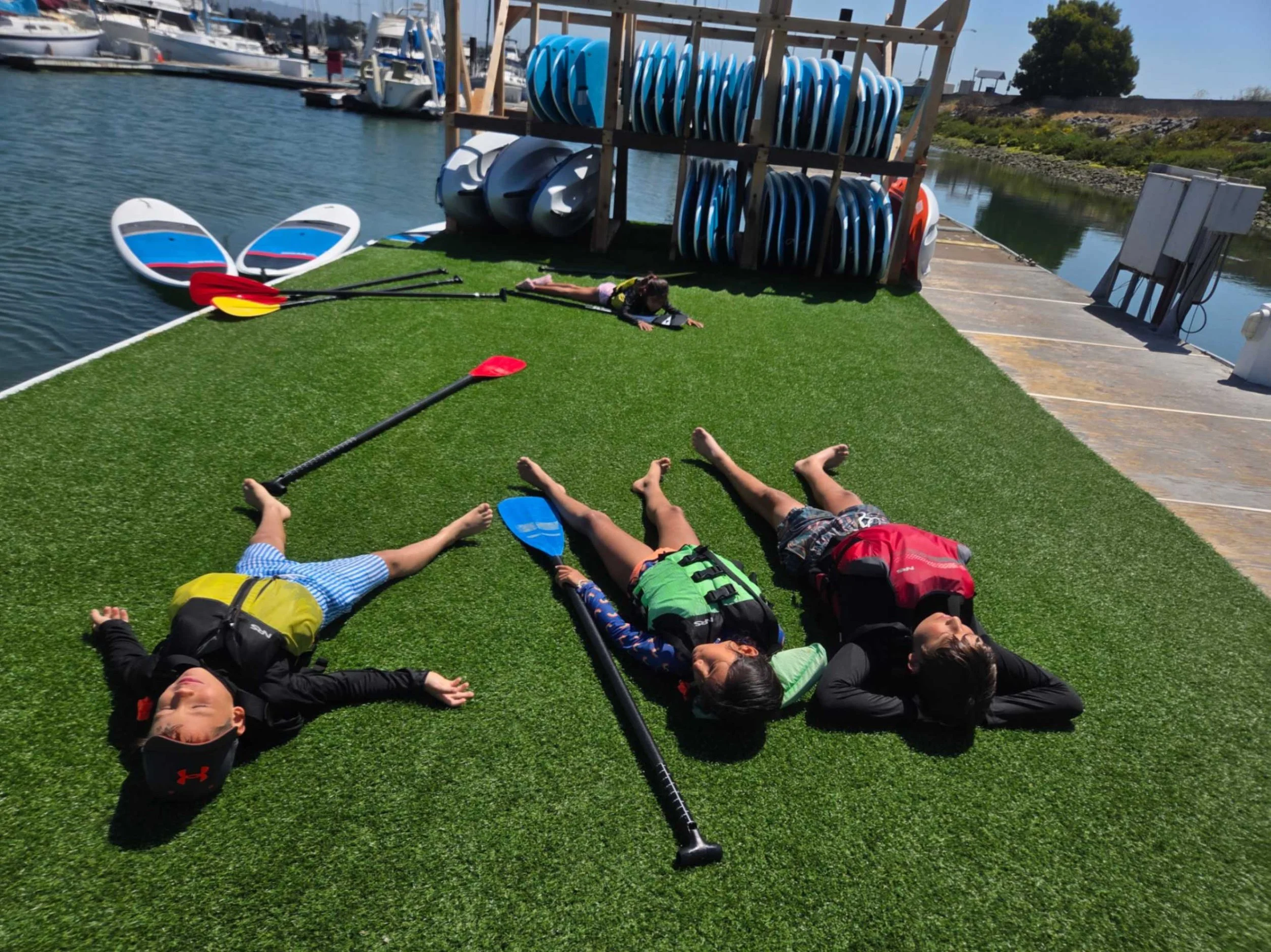 Three children lying on artificial grass next to a marina, with two paddleboards and paddles nearby, and additional paddleboards stored on a rack in the background. Having fun at Alamedas Best Summer Camp.