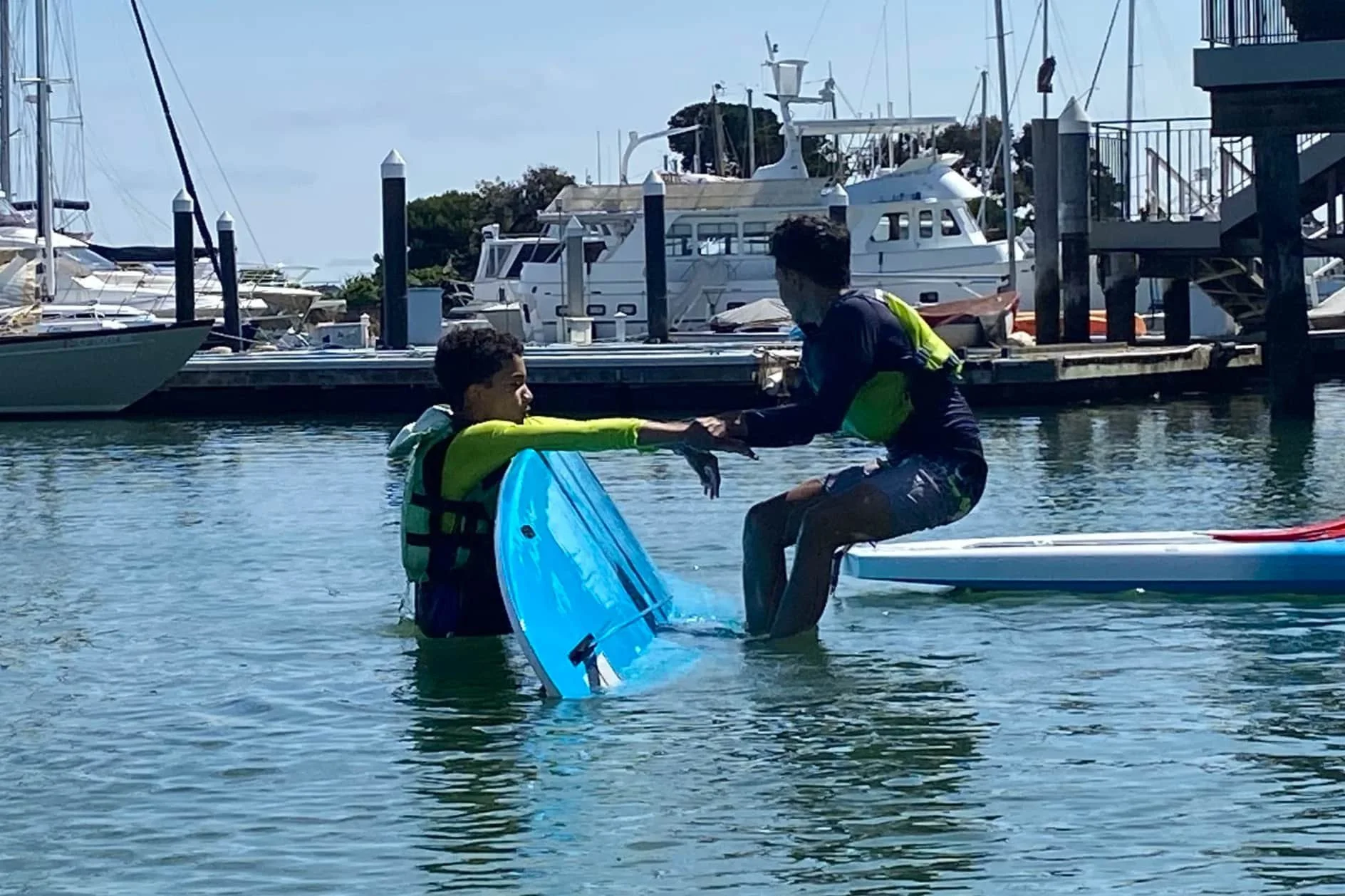 Two young boys both are wearing life jackets in the water by the dock, practicing rescue skills on a paddleboard. In the Teen Leadership Program.