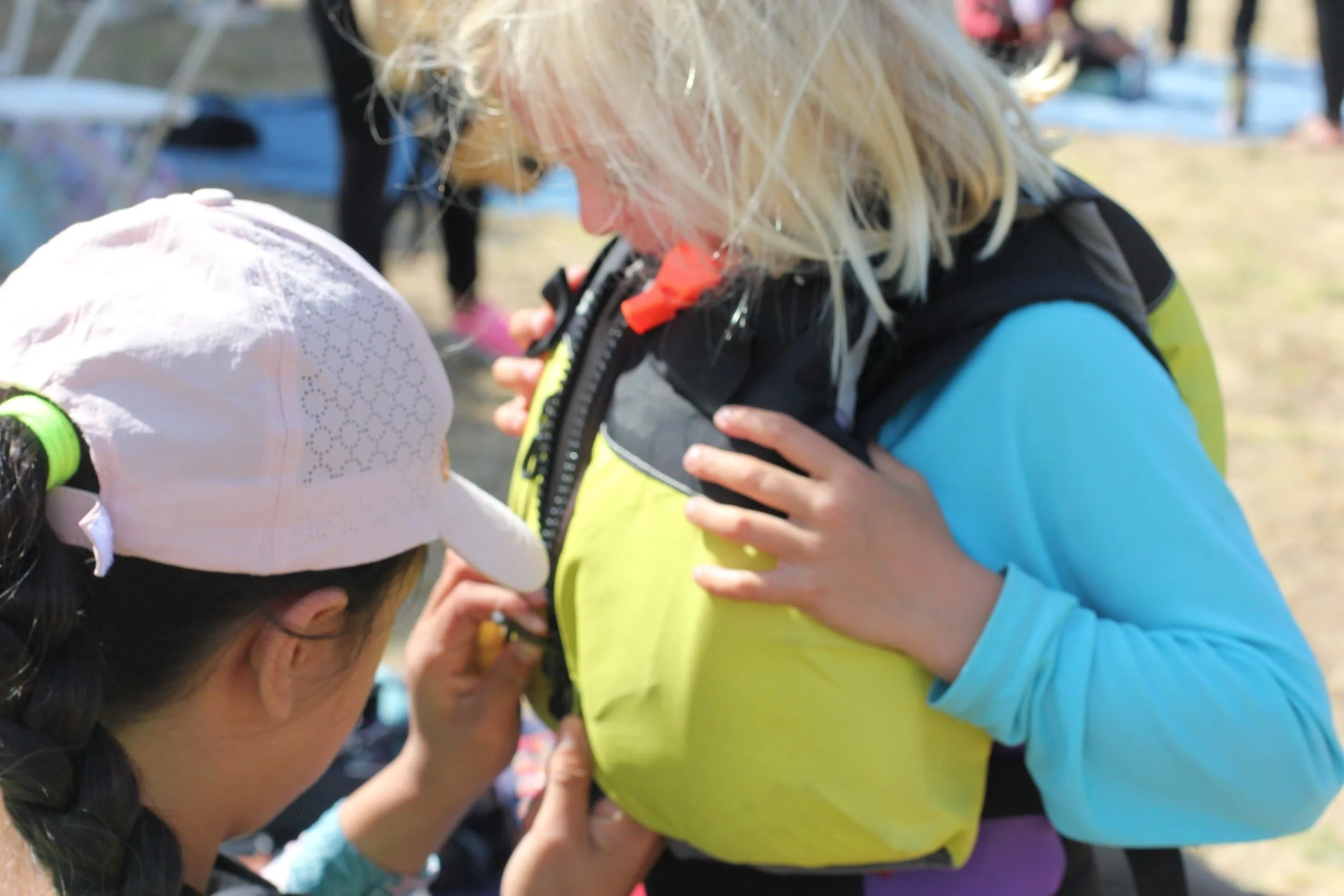 Instructor is helping a child zip up their lifejacket at summer camp while outdoors during a sunny day.