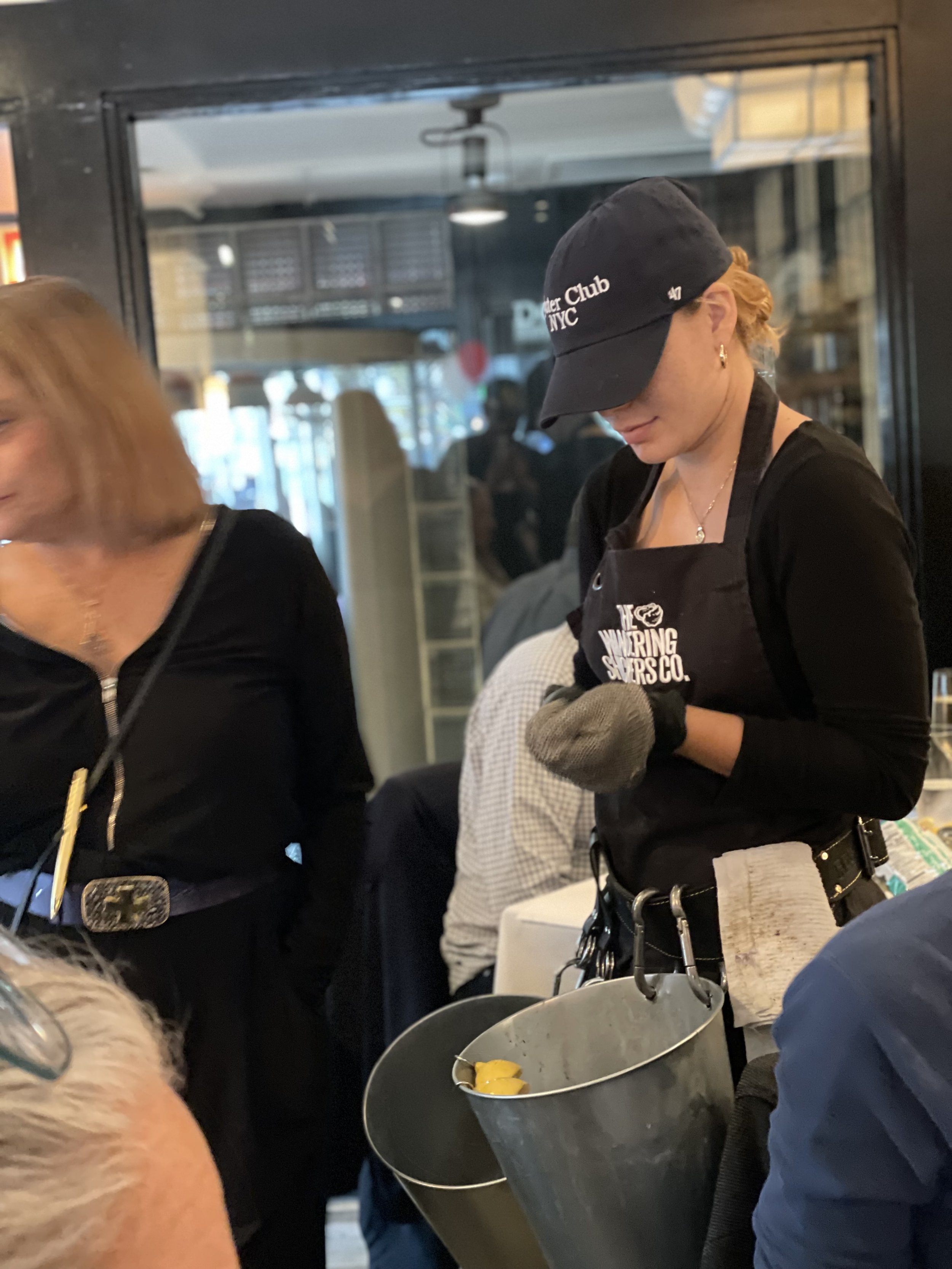 A female server wearing a black apron, gloves, and a black baseball cap serving oysters out of buckets at a restaurant or bar.
