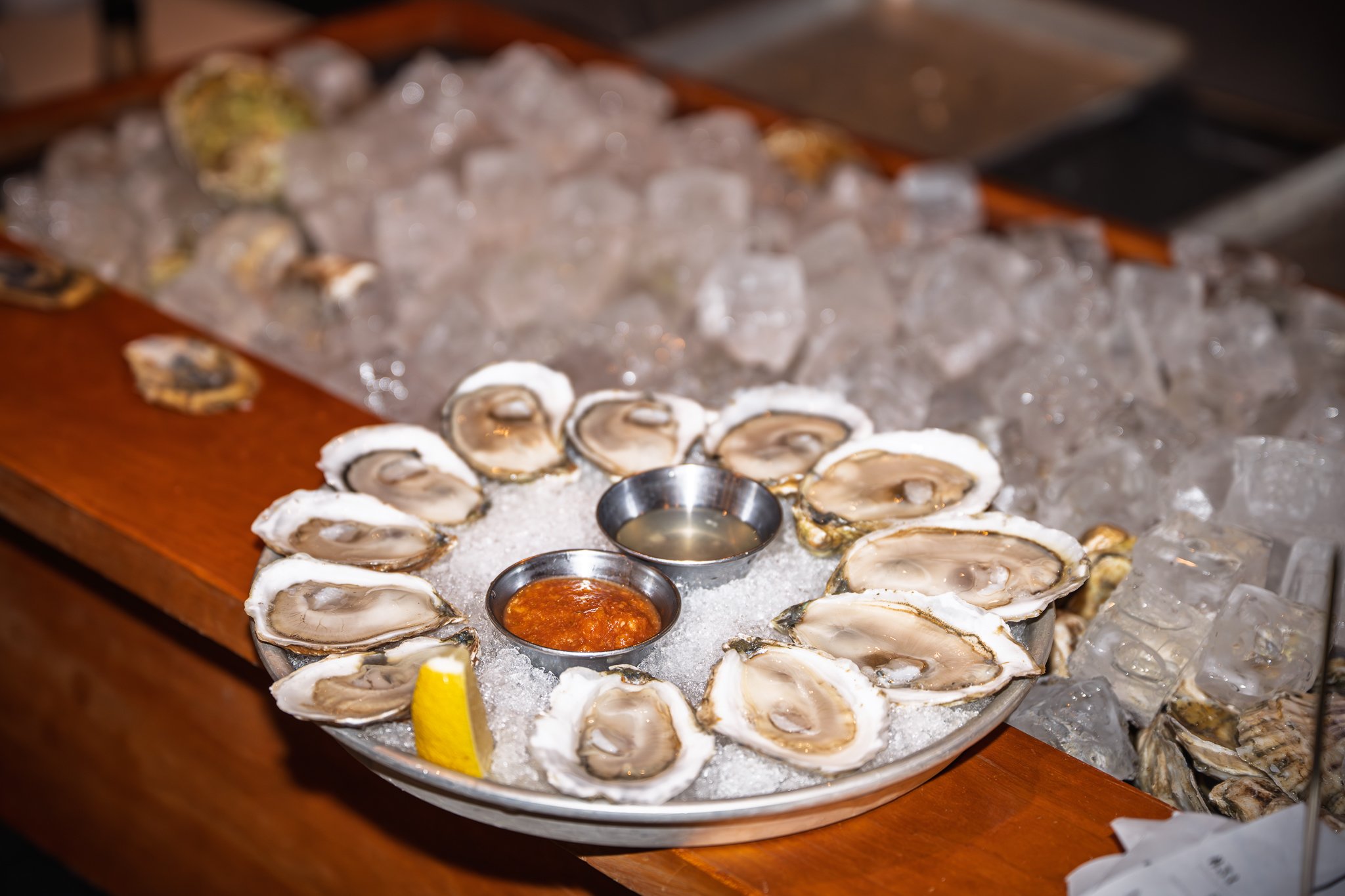 A plate of fresh oysters on ice with lemon wedges and two small bowls of cocktail sauce and mignonette, set on a bed of crushed ice.