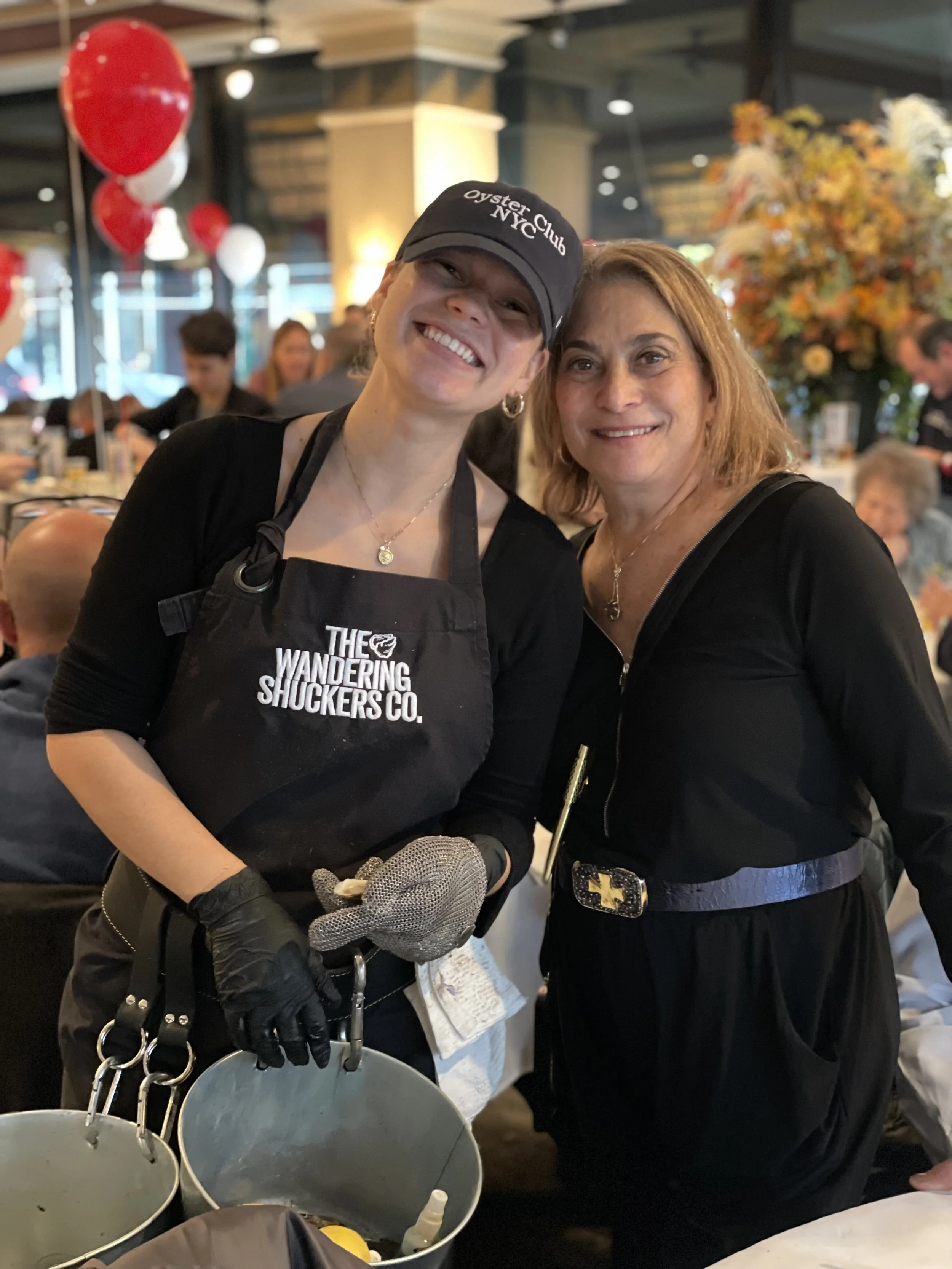 Two women smiling at a restaurant, one wearing a black apron and cap with 'Oyster Club' written on it, the other in a black top, with balloons and decorative flowers in the background.