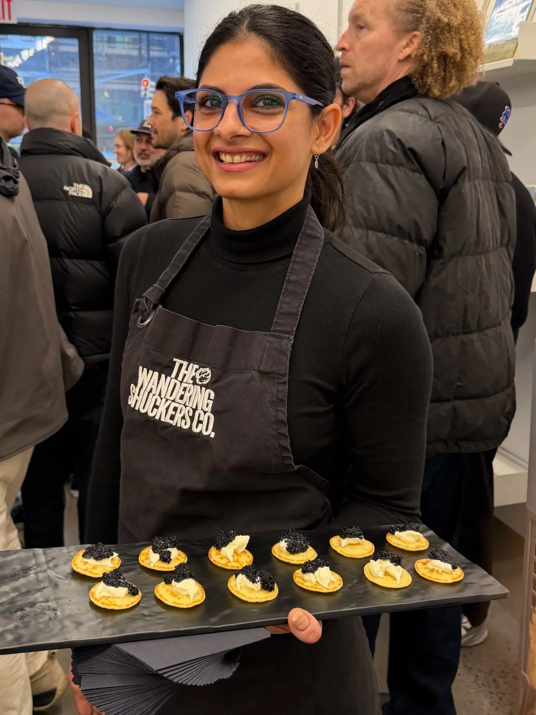 Server in a Wandering Shuckers apron holding a tray of blinis topped with caviar at a crowded event