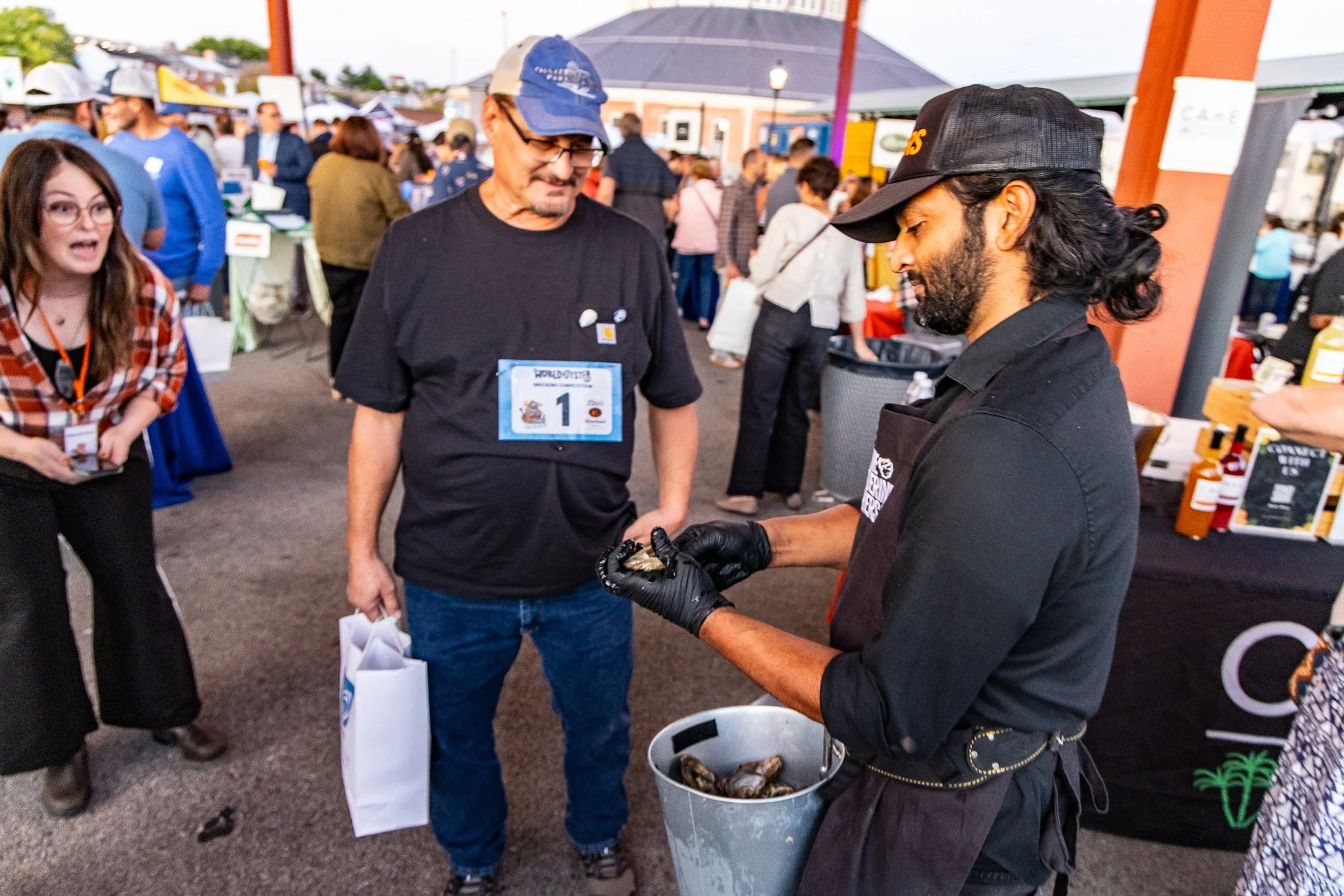 A man wearing glasses and a blue cap is looking at a chef's display of oysters at an outdoor event, surrounded by crowds and vendor booths.