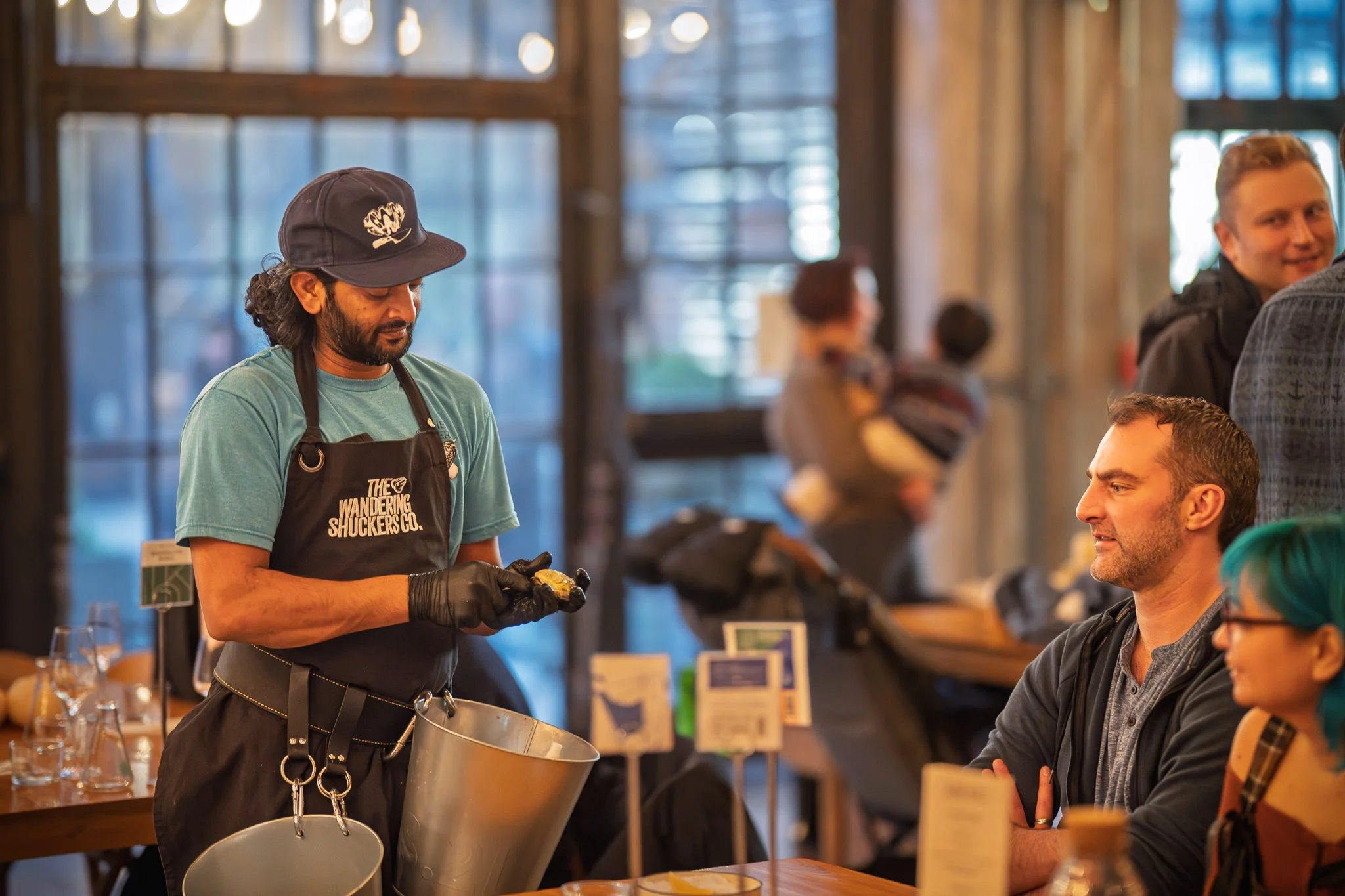 A man in a blue t-shirt, black apron with text, and black gloves is preparing food at a wooden counter inside a rustic-style market or cafe. Several customers are seated at the counter, observing him, including a man in a gray hoodie and a woman with teal hair and glasses. In the background, a woman holds a child, and other people are seen walking or standing in the space with large windows and wooden decor.