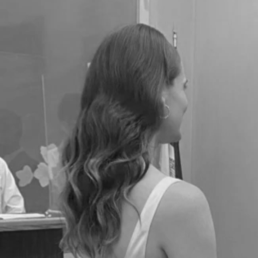 Side view of a woman with wavy brown hair wearing a white dress, standing indoors near a cubicle and a glass window.
