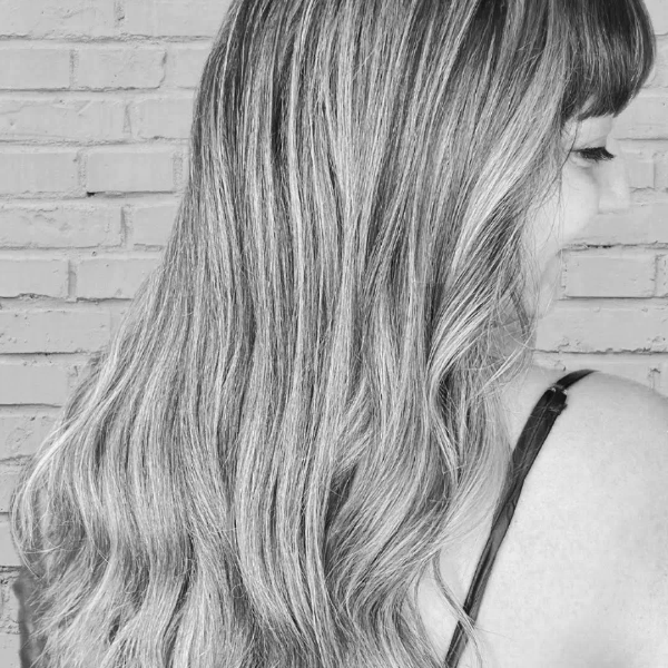 Side view of a woman with long, wavy blonde hair with darker roots and highlights, standing against a white brick wall.