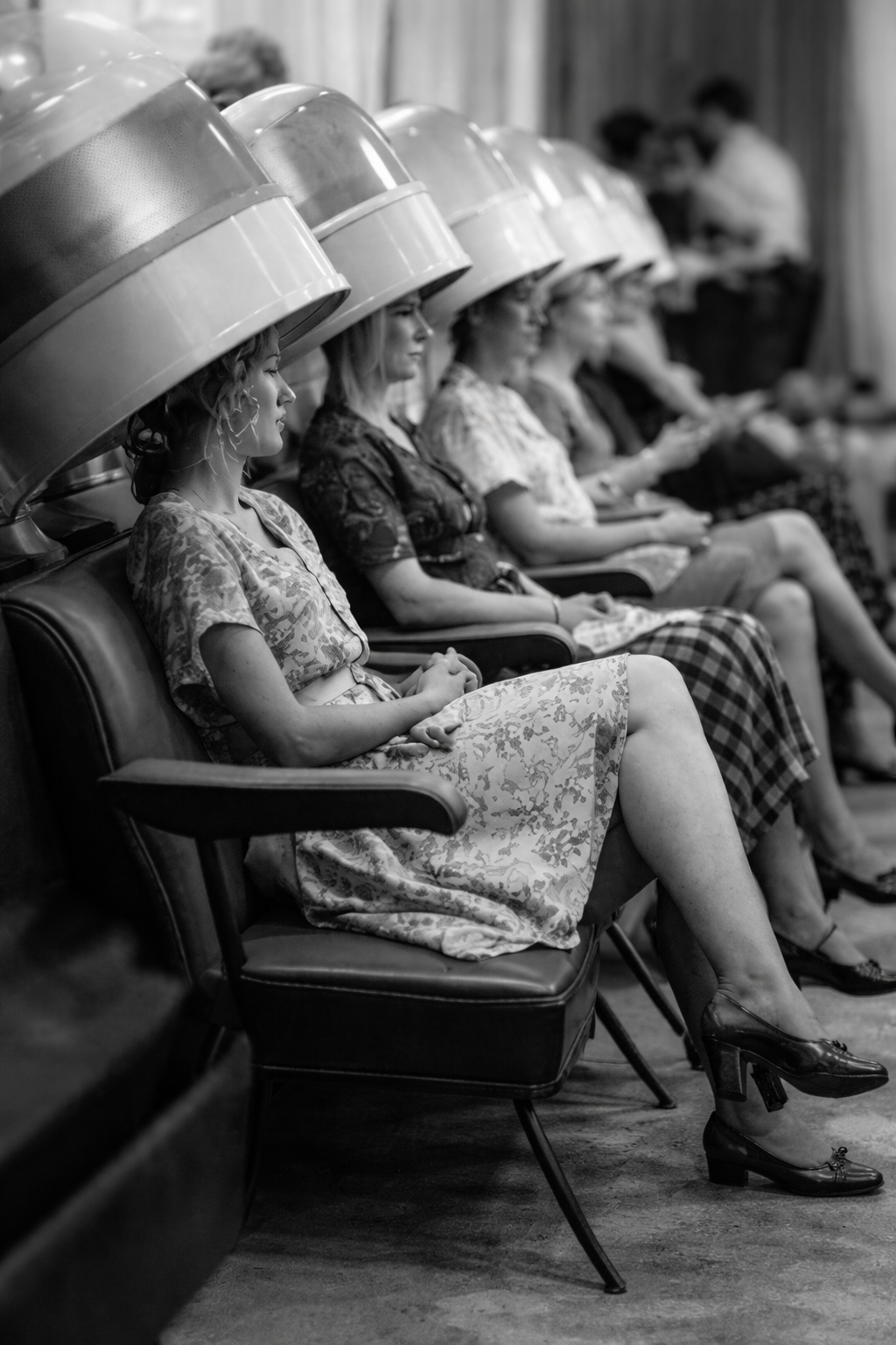 Women sitting under hair dryers in a salon, waiting for their hair to dry, in black and white.