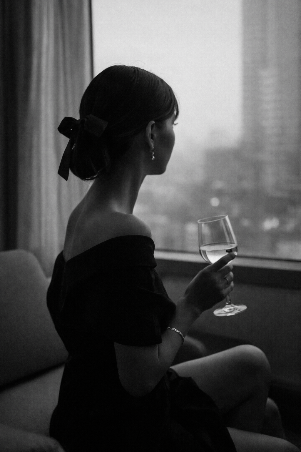 A black and white photo of a woman sitting on a couch, looking out a window while holding a glass of wine, dressed in an off-the-shoulder top and wearing earrings and a bracelet.