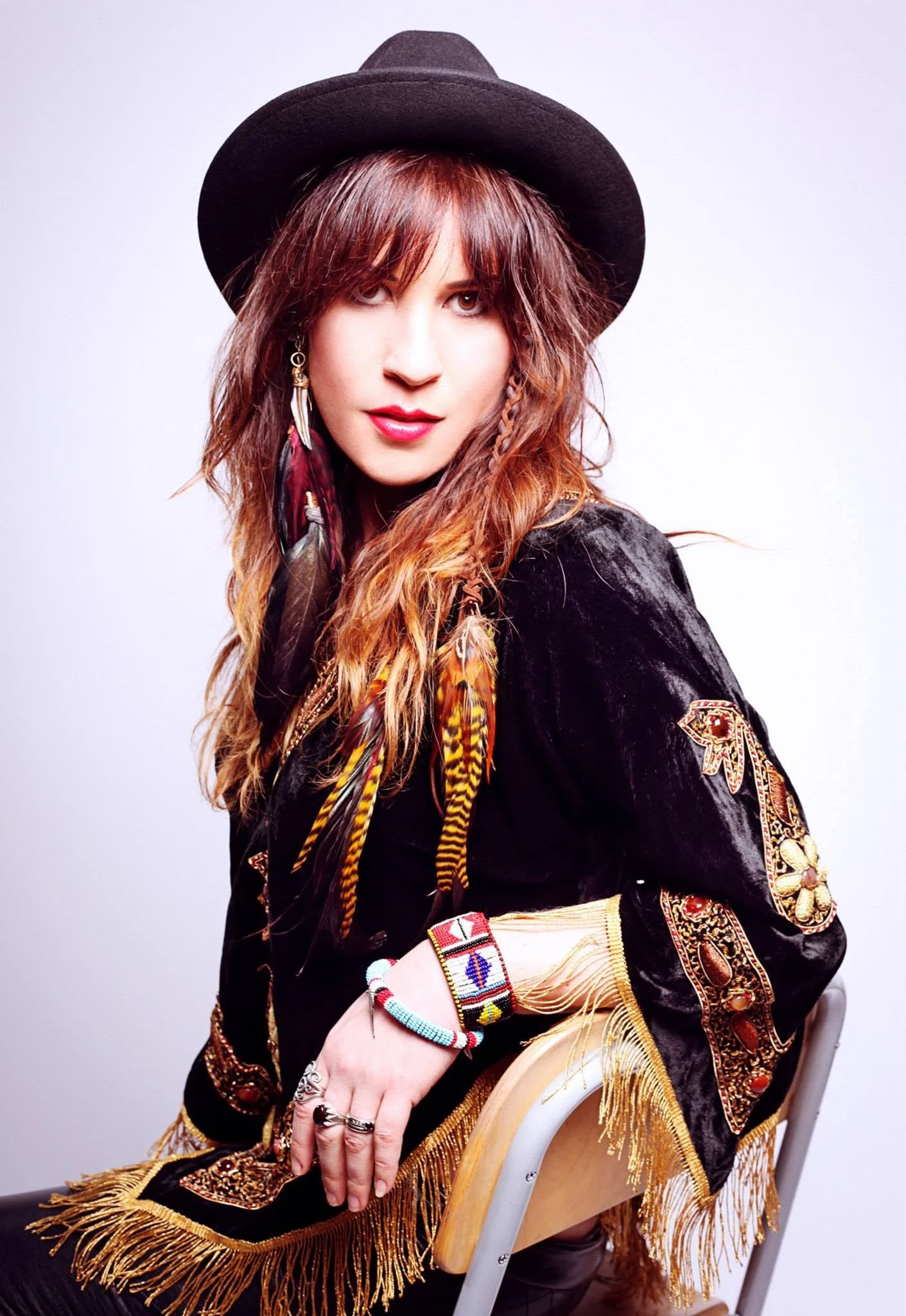 Woman wearing a black wide-brim hat with a feathered headband, a black jacket with embroidered and beaded details, sitting on a chair with beaded jewelry and a fringed shawl.