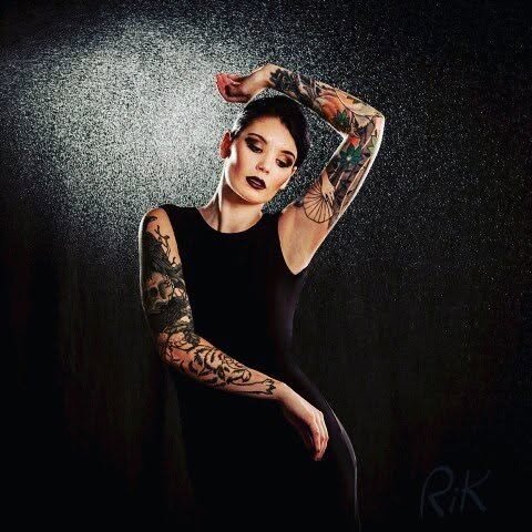 A woman with tattooed arms in a black dress, with wet hair, standing against a dark background, and holding her arm above her head.