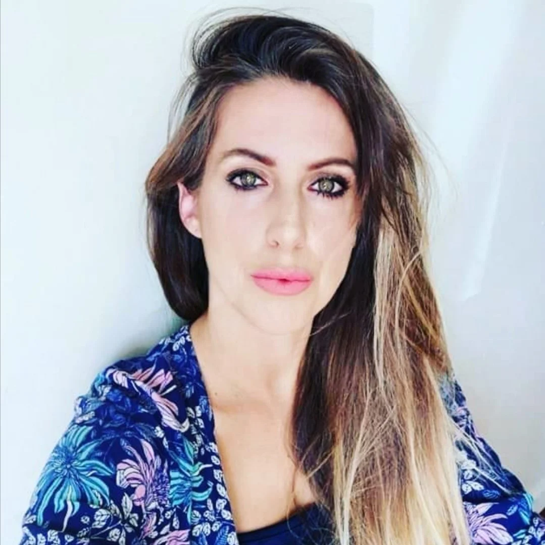 Kaz Elizabeth founder of Glamour Tribe Makeup Artistry A woman with long, wavy hair and makeup, wearing a blue patterned top, posing in front of a light background.