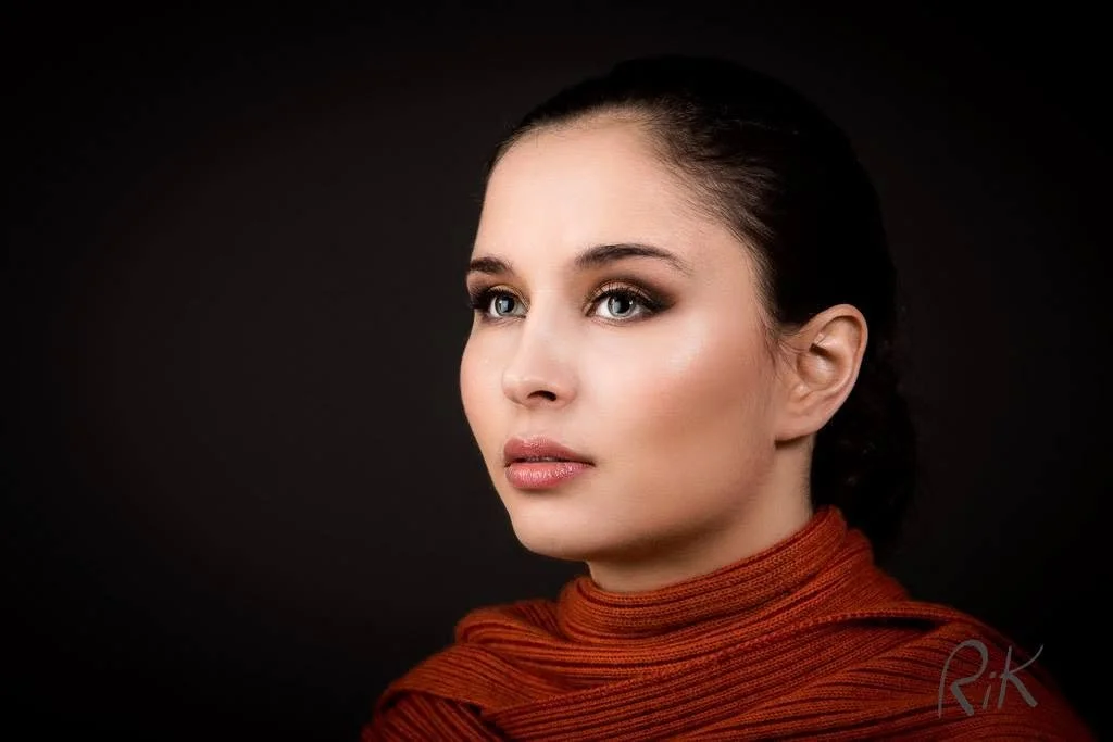 A portrait of a young woman with blue eyes and dark hair styled back, wearing a rust-colored turtleneck sweater against a dark background.