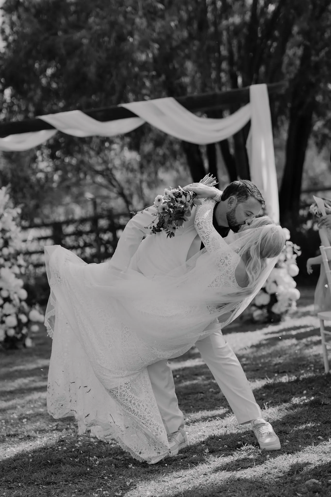 Kaz Elizabeth makeup artist galmour tribe makeup artistry team  man in formal attire leaning down to kiss a woman in a wedding dress during an outdoor wedding ceremony, with draped fabric and trees in the background.