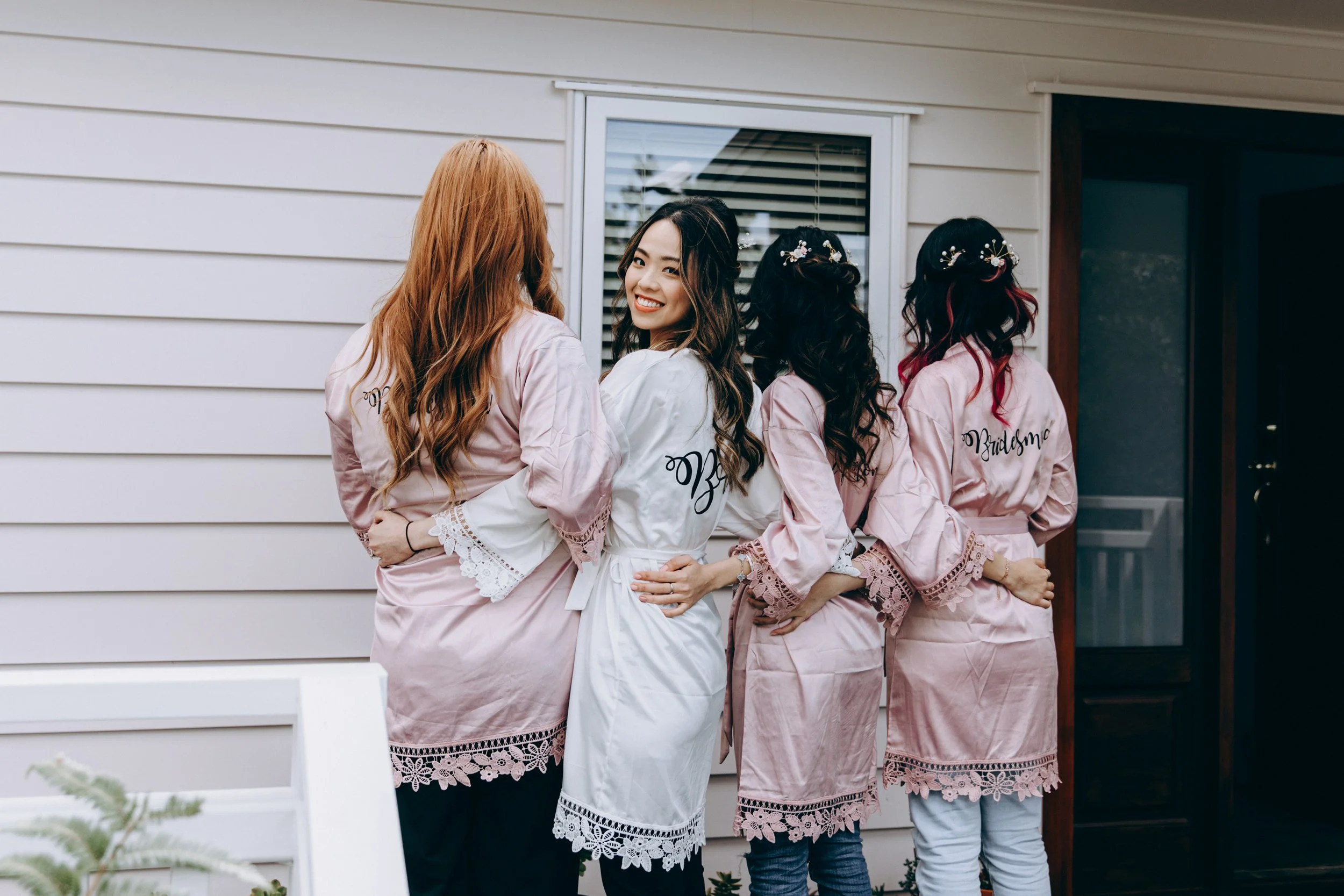 Four women dressed in satin robes with lace trim, embracing each other against a house wall. One woman is smiling at the camera, and the others have their backs to the camera, with one wearing a robe with 'Bridesmaid' written on it and floral hair accessories.