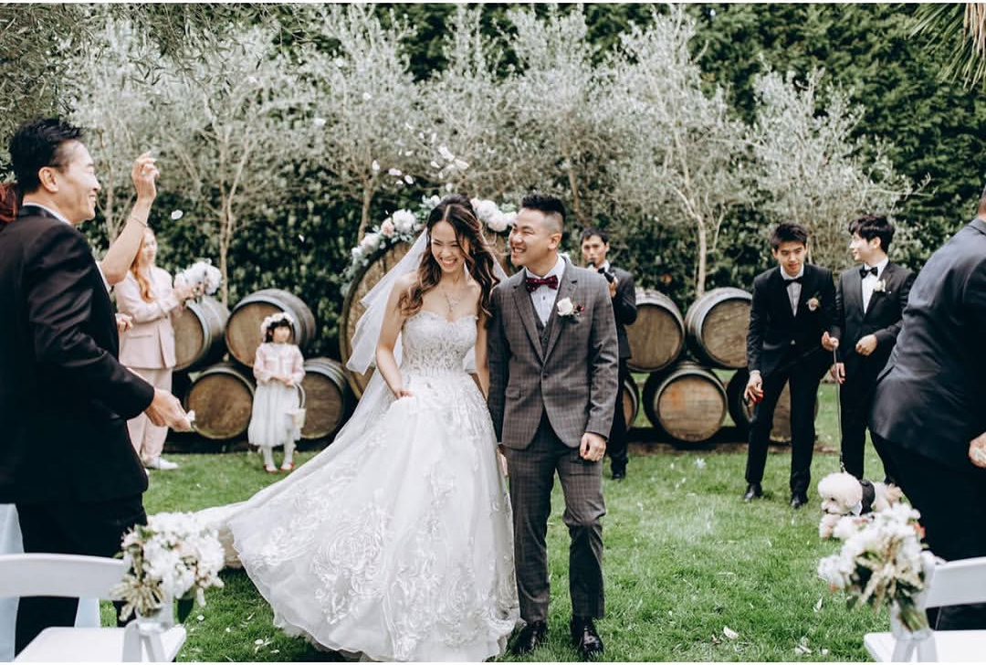 A bride and groom smiling and walking together at an outdoor wedding ceremony, with family and friends surrounding them.