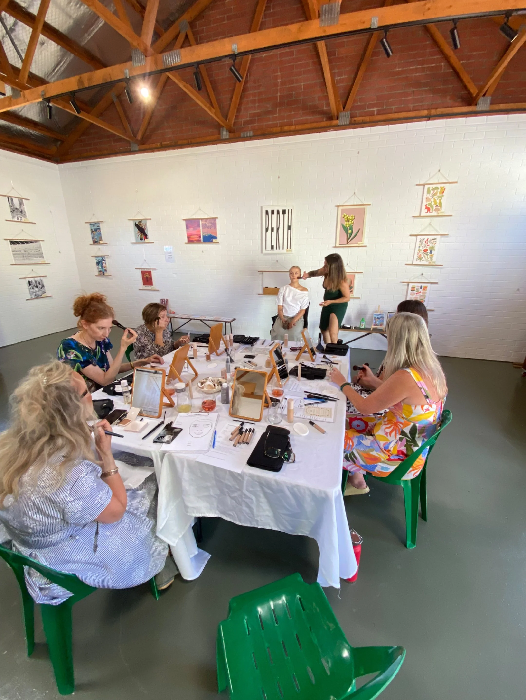 A group of women gathered around a table doing makeup and beauty activities at an indoor event space with white brick walls decorated with art and a sign that says 'PERTH'. Some women are sitting at the table using makeup and tools, while a woman standing appears to be performing a demonstration or makeup application on another woman.