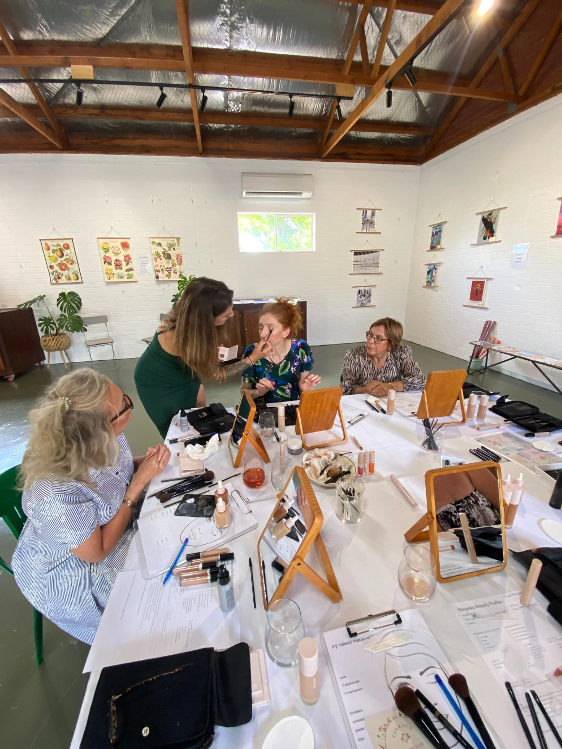 People sitting at a long table with makeup products, brushes, and papers, with several art prints hanging on white brick walls. One woman is applying makeup to another, while others observe or prepare. The room has a high ceiling with insulation and track lighting.