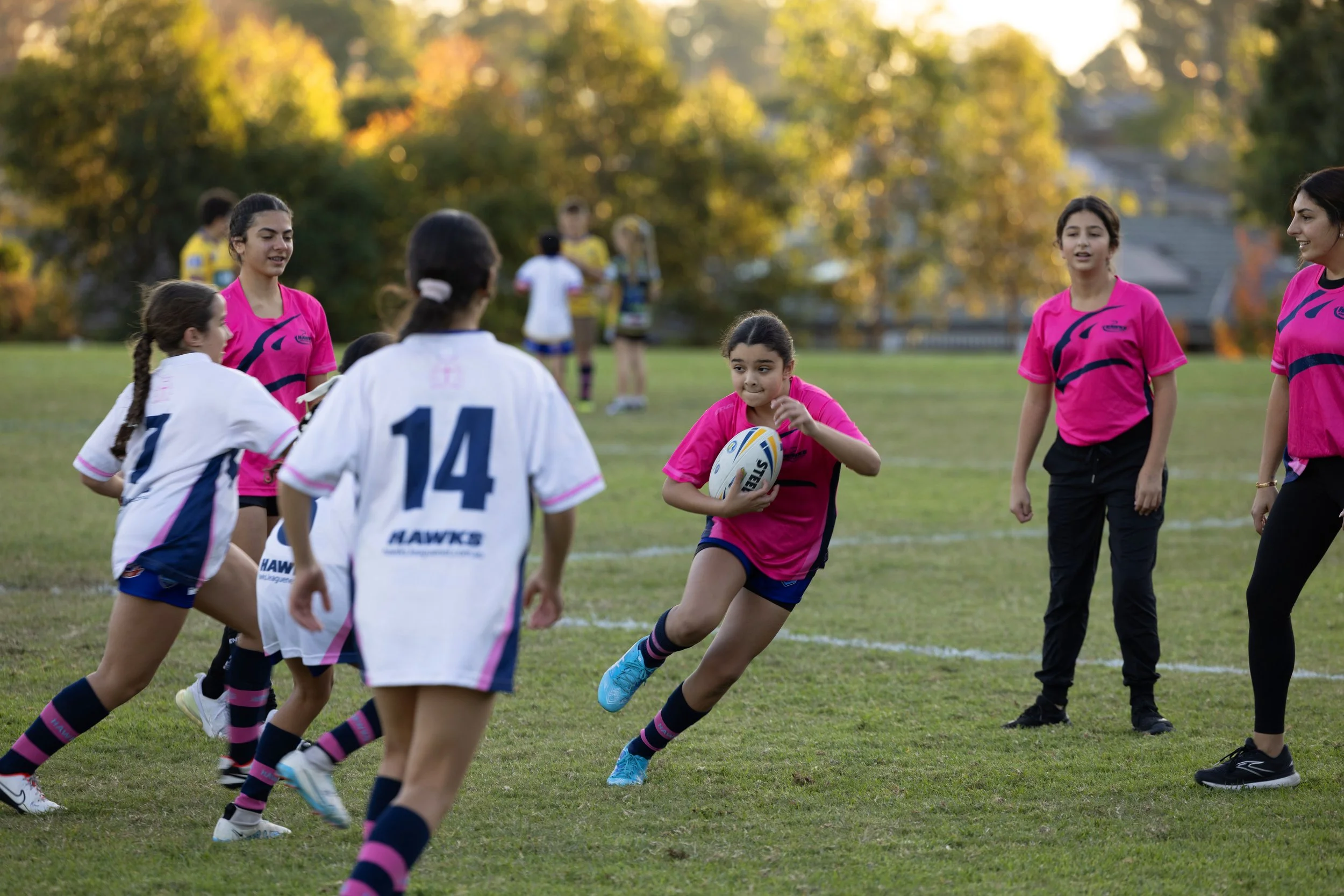 Girls playing rugby on a grassy field during the daytime. One girl in a pink shirt is running with a rugby ball, while other girls and women are standing or moving nearby, watching or participating in the game.