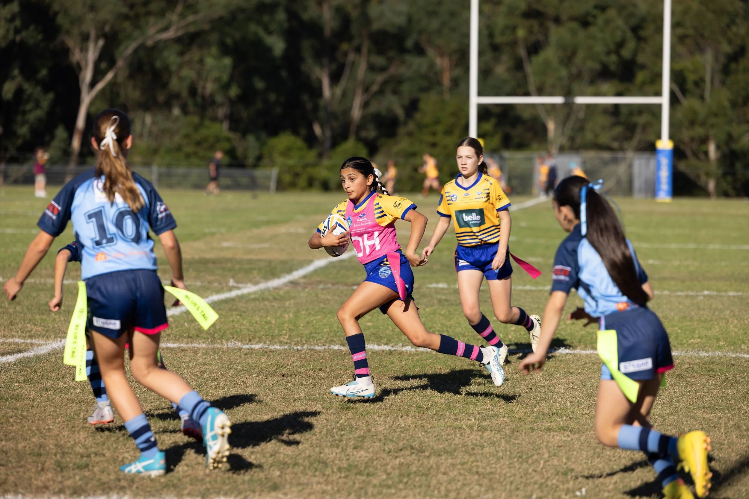 Girls playing rugby on a field, one girl running with a rugby ball, surrounded by opponents in blue jerseys. Background of goal post and trees.