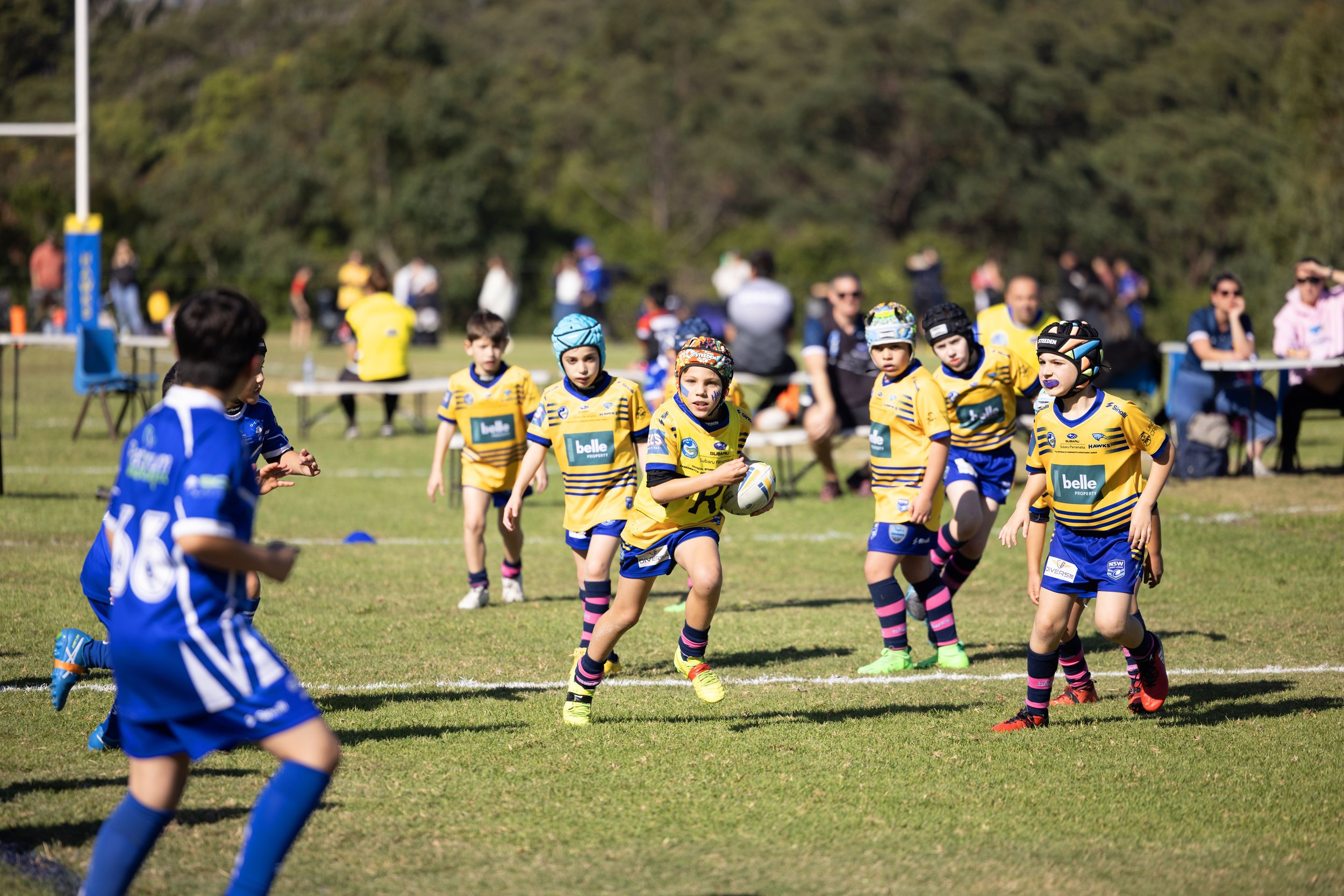 Children playing rugby on a grassy field with spectators in the background