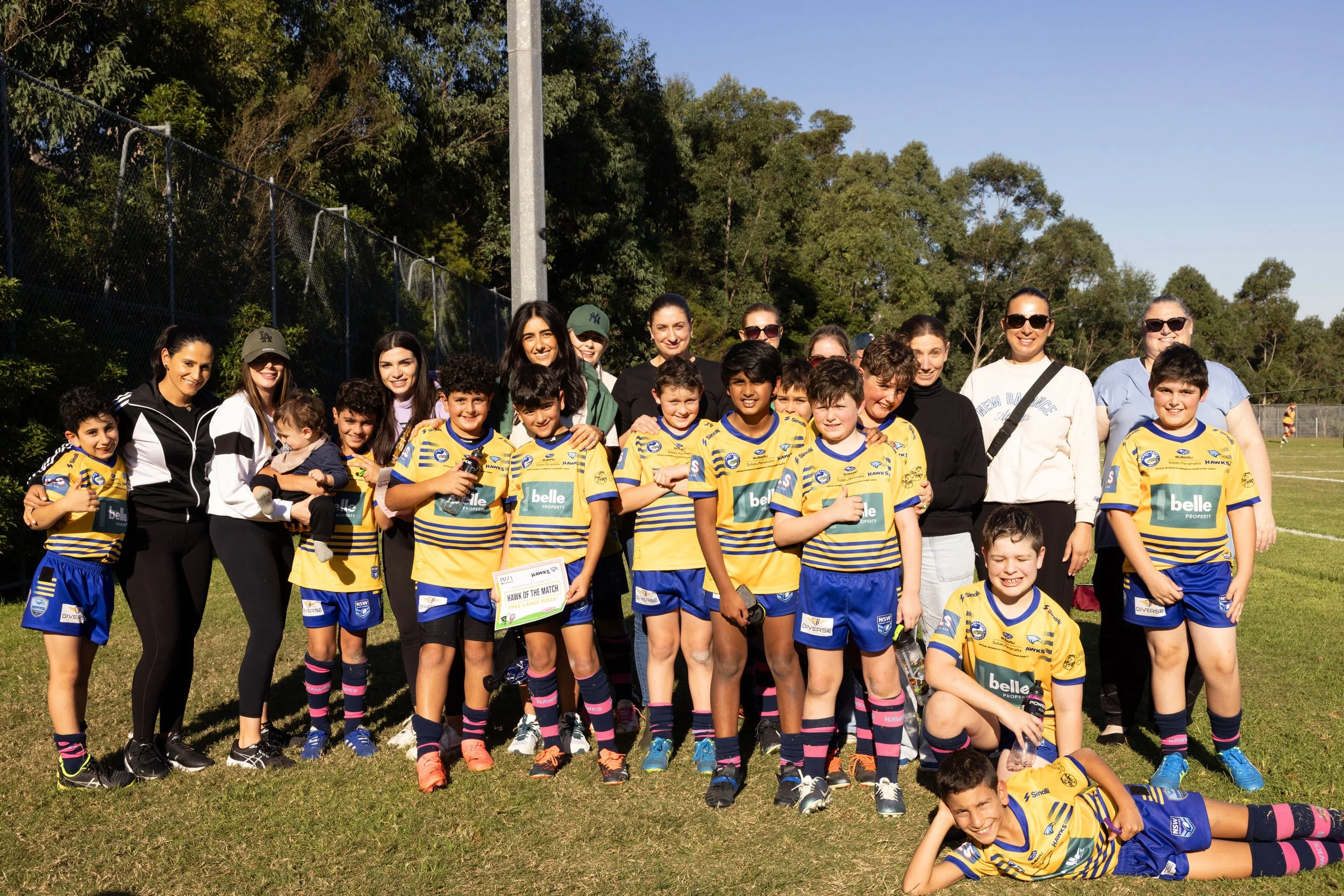 A group of children and adults posing on a grassy field after a youth sports match. The children are wearing yellow and blue sports uniforms, some holding a 
