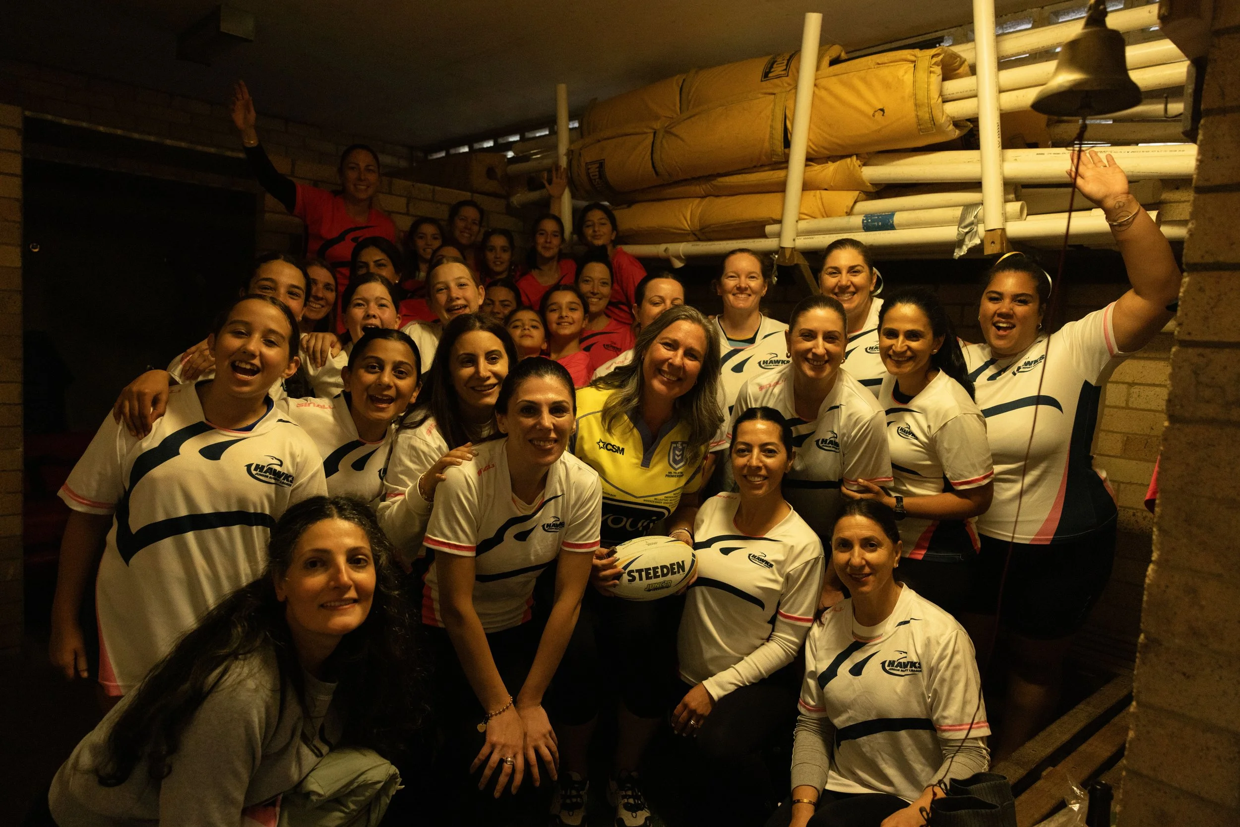 Group of women in sports uniforms, some holding a rugby ball, taking a group photo indoors with brick walls and sports equipment in the background.