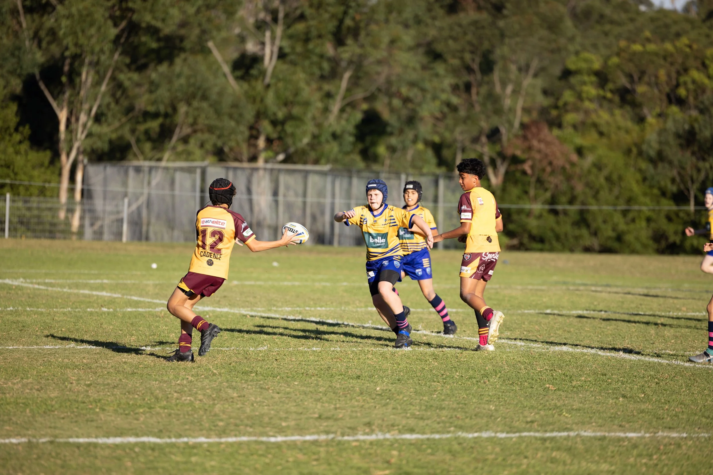 Children playing rugby on a grassy field with trees in the background.