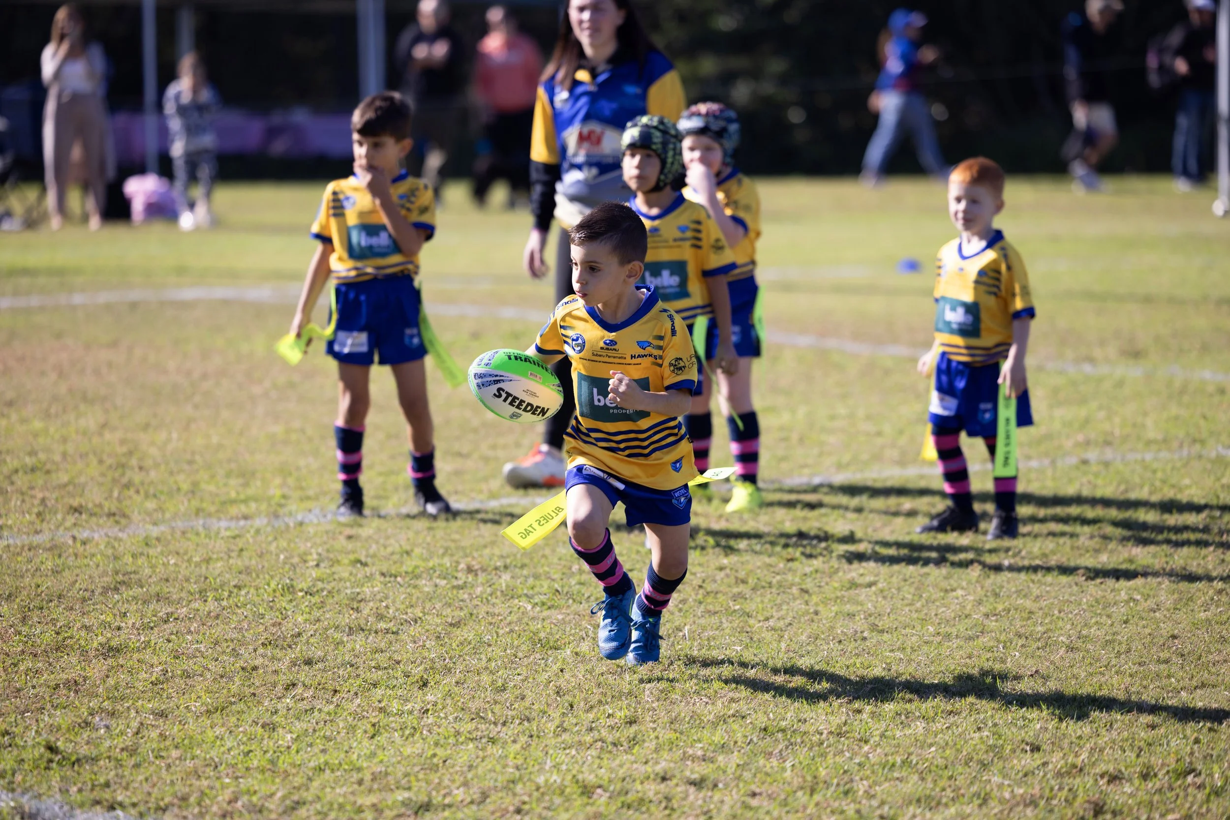 Young children in yellow and blue rugby uniforms playing on a grassy field during daytime, with some children stopping and others running, a young boy reaching for a rugby ball in the foreground.
