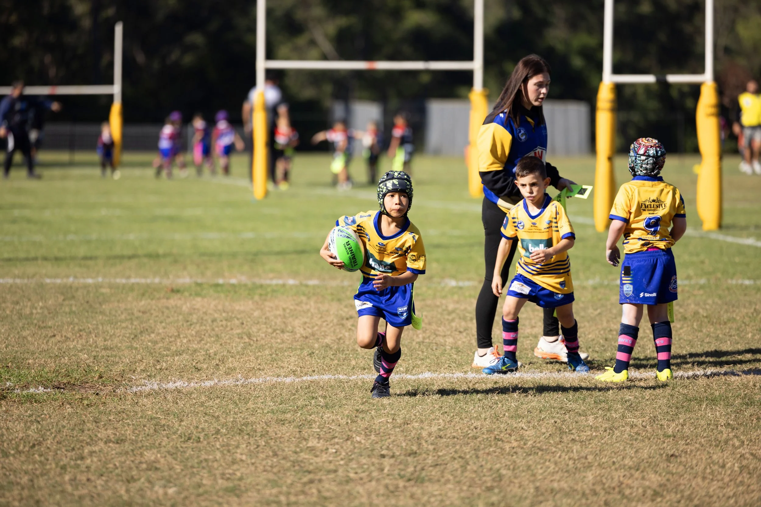 Children playing rugby on a field with a coach or referee nearby, and other players visible in the background.