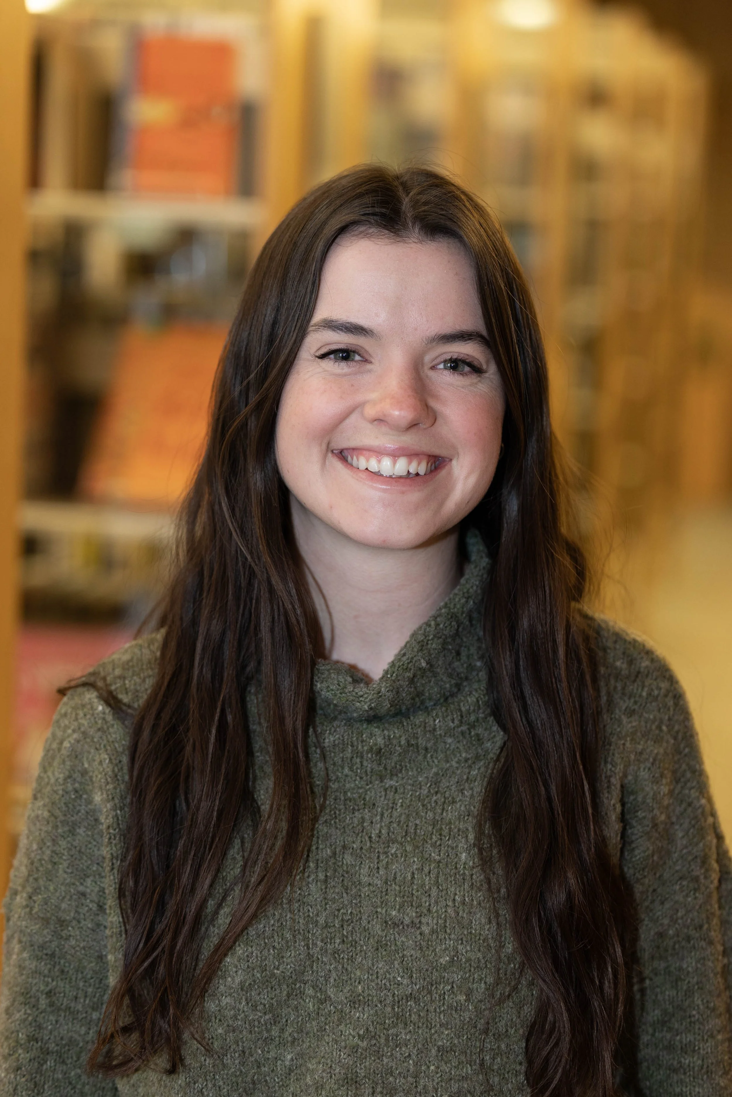 A young woman with long brown hair and blue eyes smiling in front of a blurred background of a library with bookshelves.