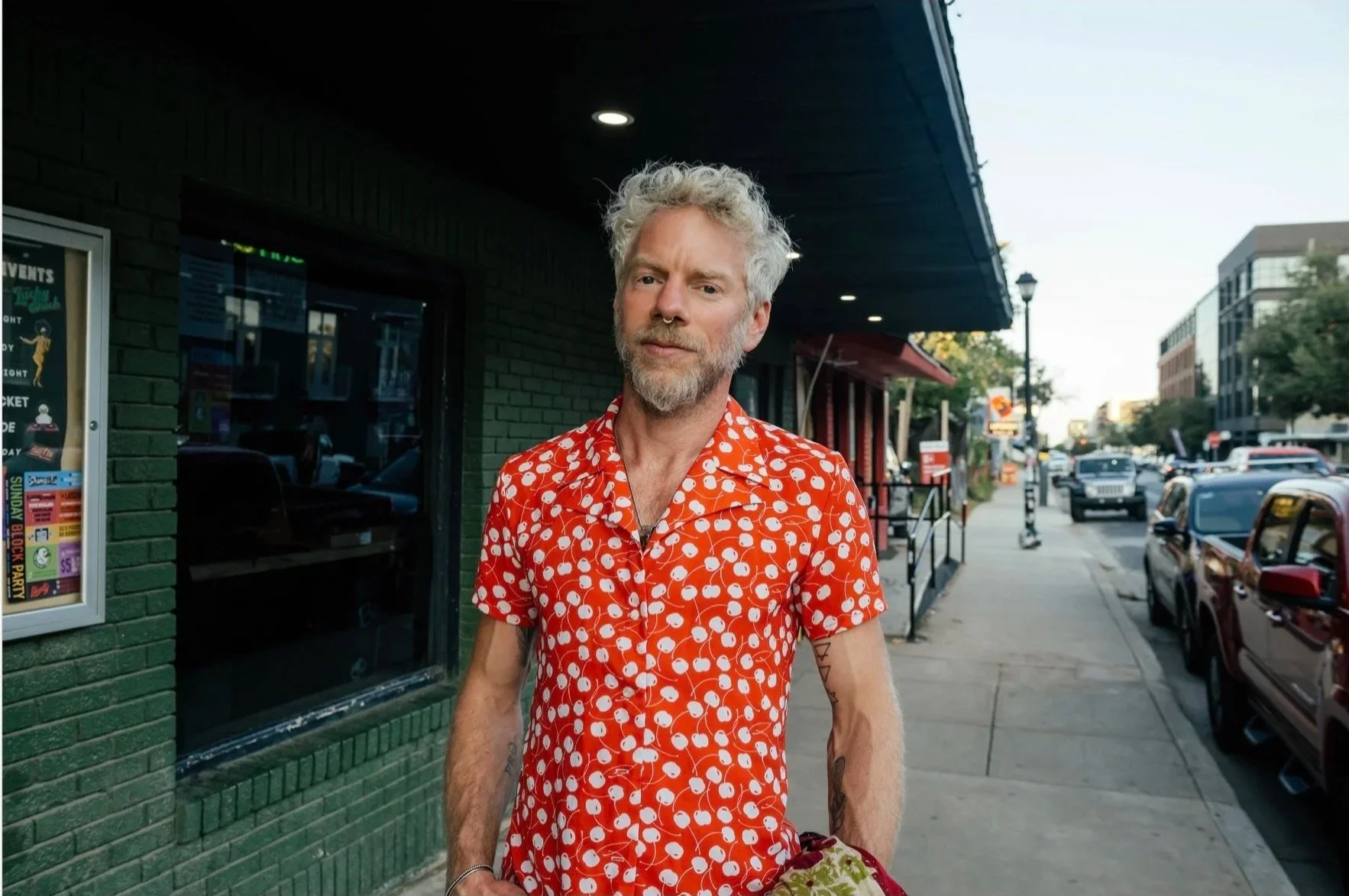 A man with curly, light-colored hair and a beard stands on a city sidewalk wearing a red shirt with a white cherry pattern, holding a floral cloth, with cars and buildings visible in the background.