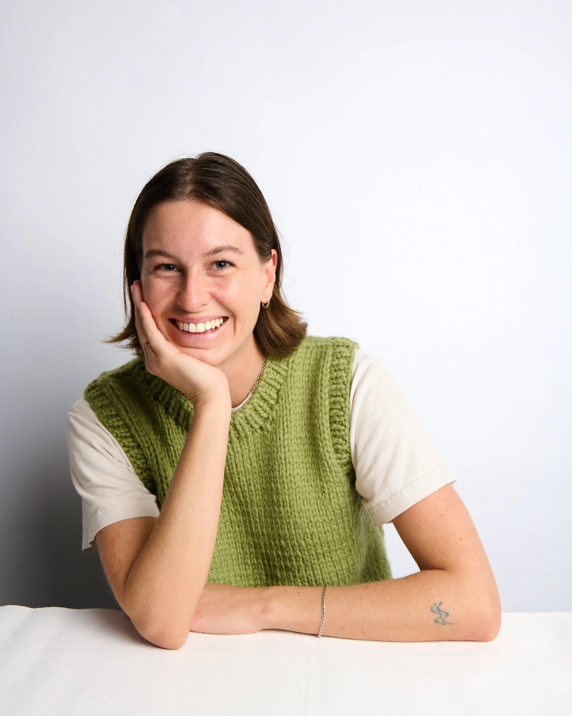 Young woman smiling, wearing a green knitted vest over a white shirt, resting her head on her hand, with a small tattoo on her forearm, sitting at a white table in front of a white background.