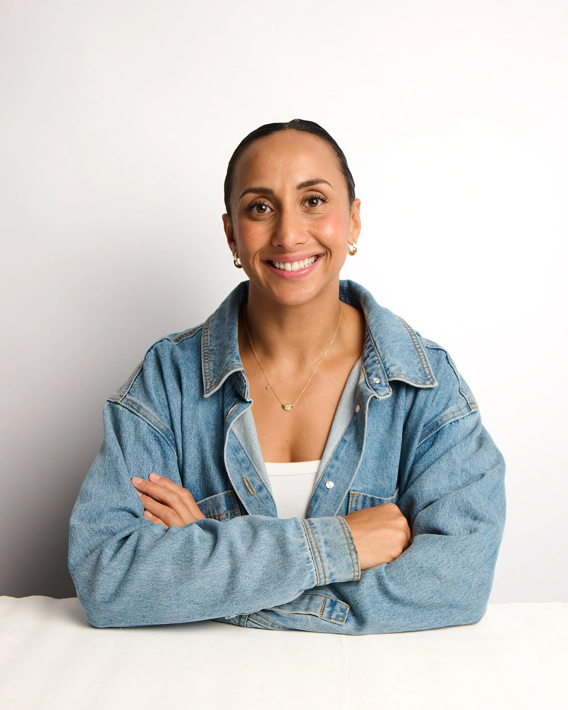 Portrait of a woman with dark hair, smiling and crossing her arms, wearing a denim jacket and a white top, against a white background.