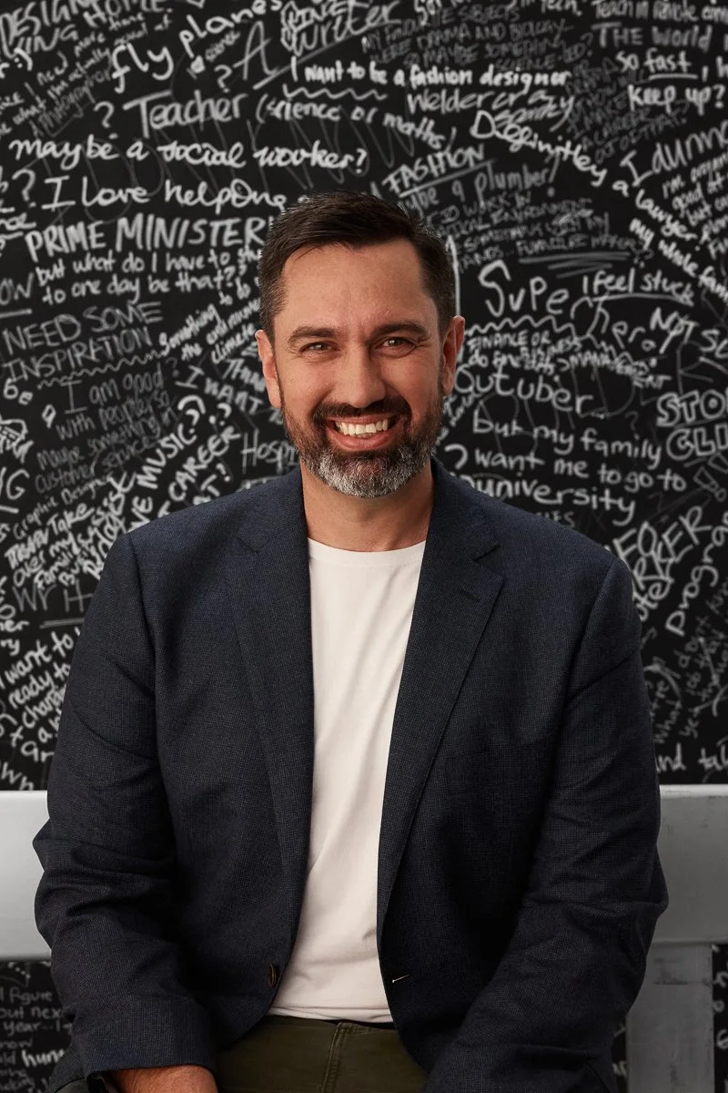 A man sitting on a metal bench seat, in a studio. He is smiling. There is a paper backdrop behind him covered in writing done by school students who are thinking about what they want to be when they leave school.