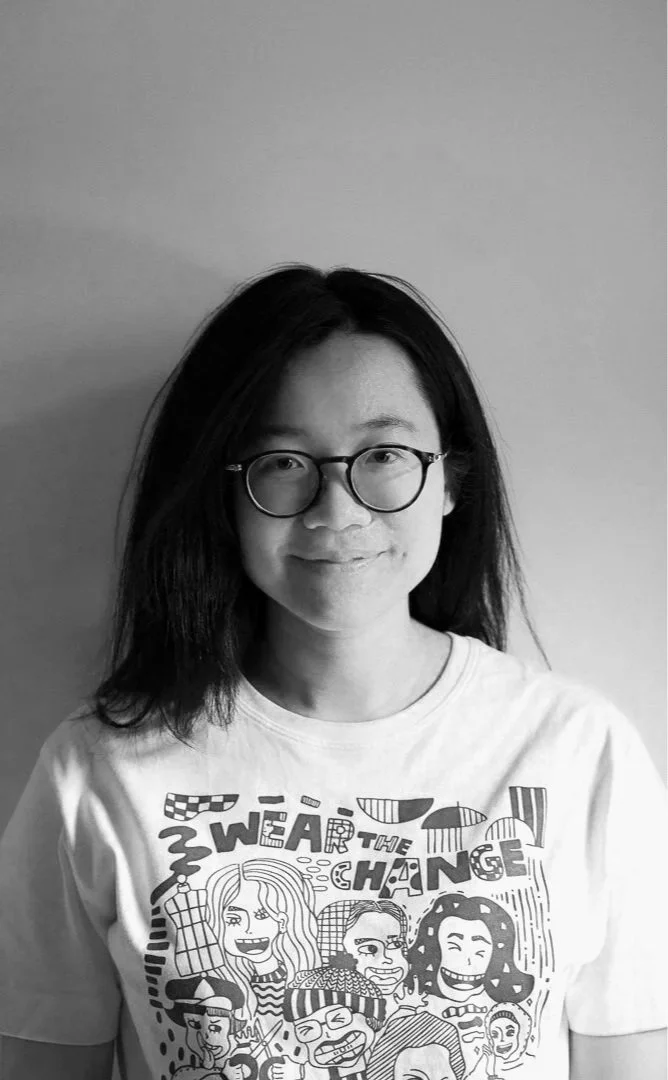 A young woman with black hair, glasses, and a white graphic t-shirt standing against a plain wall.