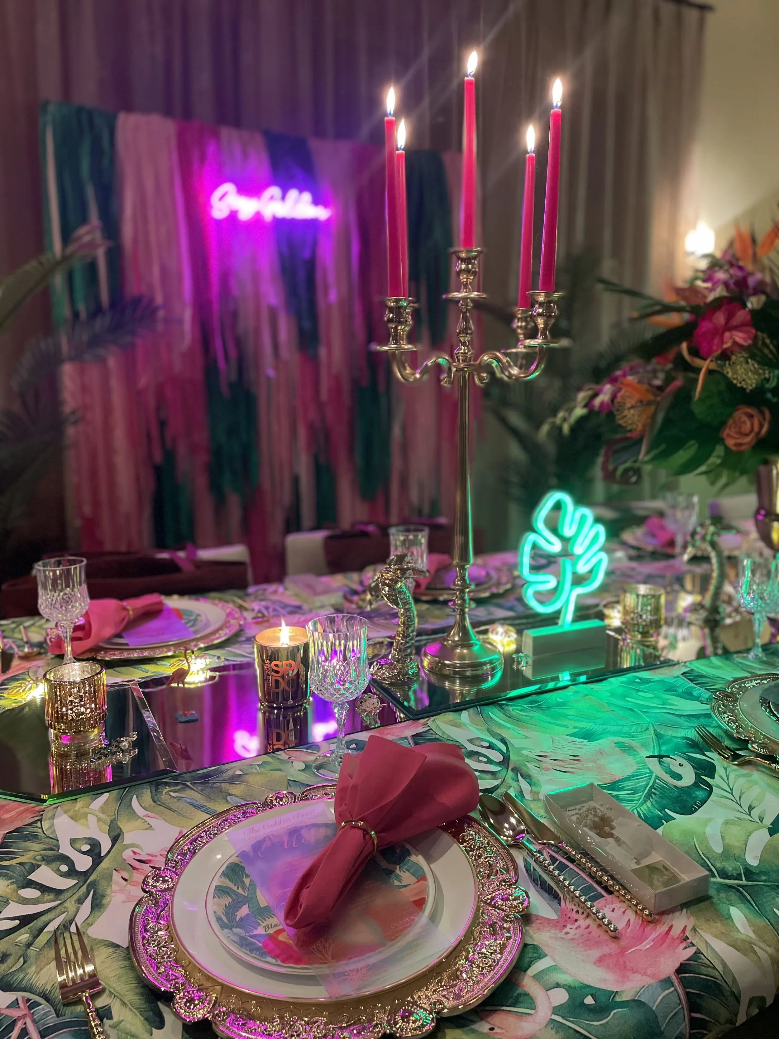 Custom Dinner design. A decorated tropical-themed party table with floral tablecloth, gold and pink plates, pink napkins, crystal glasses, candles, and pink candles in a silver candelabrum. Neon signs and a floral arrangement are in the background.