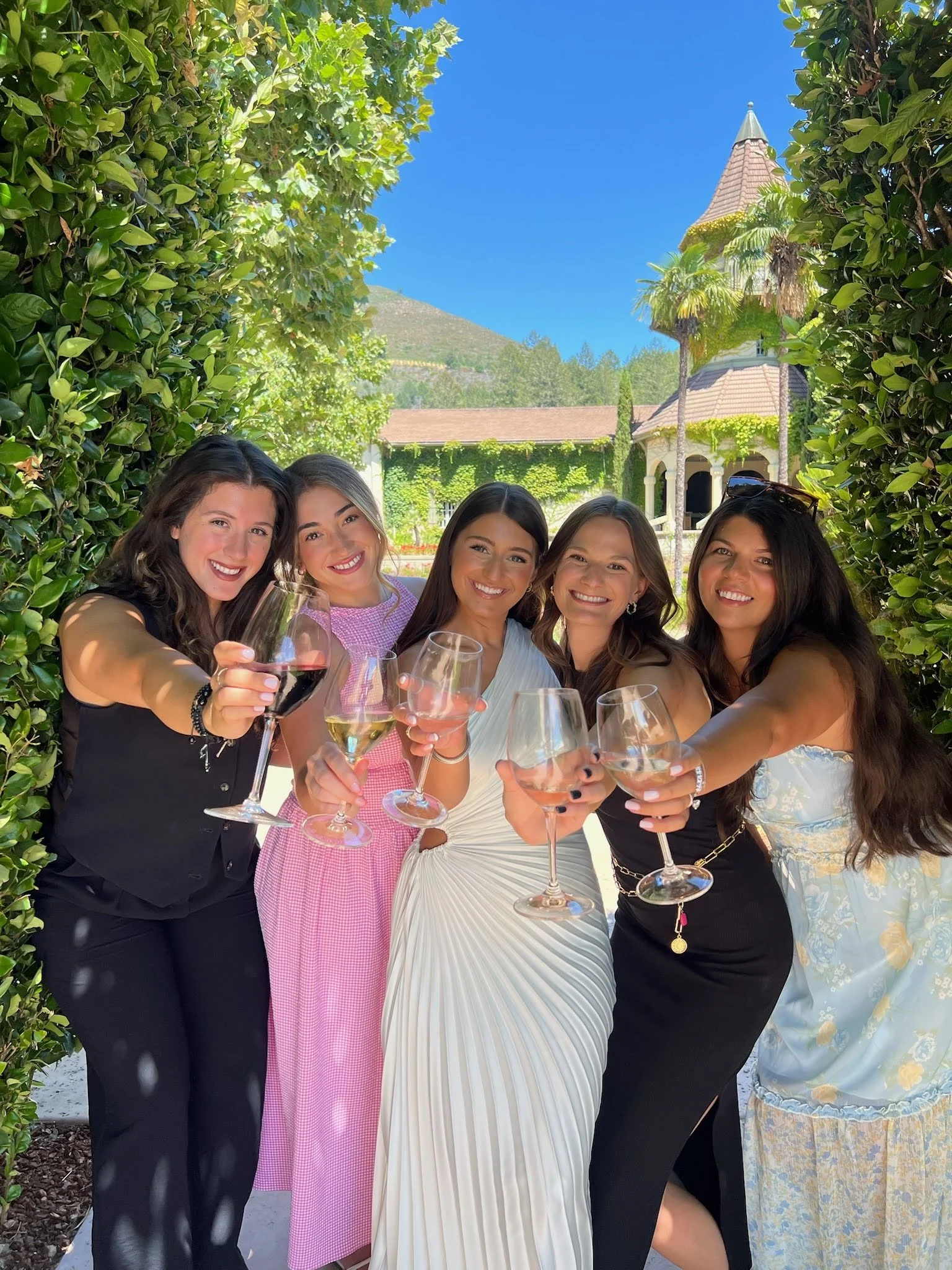 Five women clinking wine glasses outdoors with a lush green hedge and a building with a turret in the background on a sunny day.